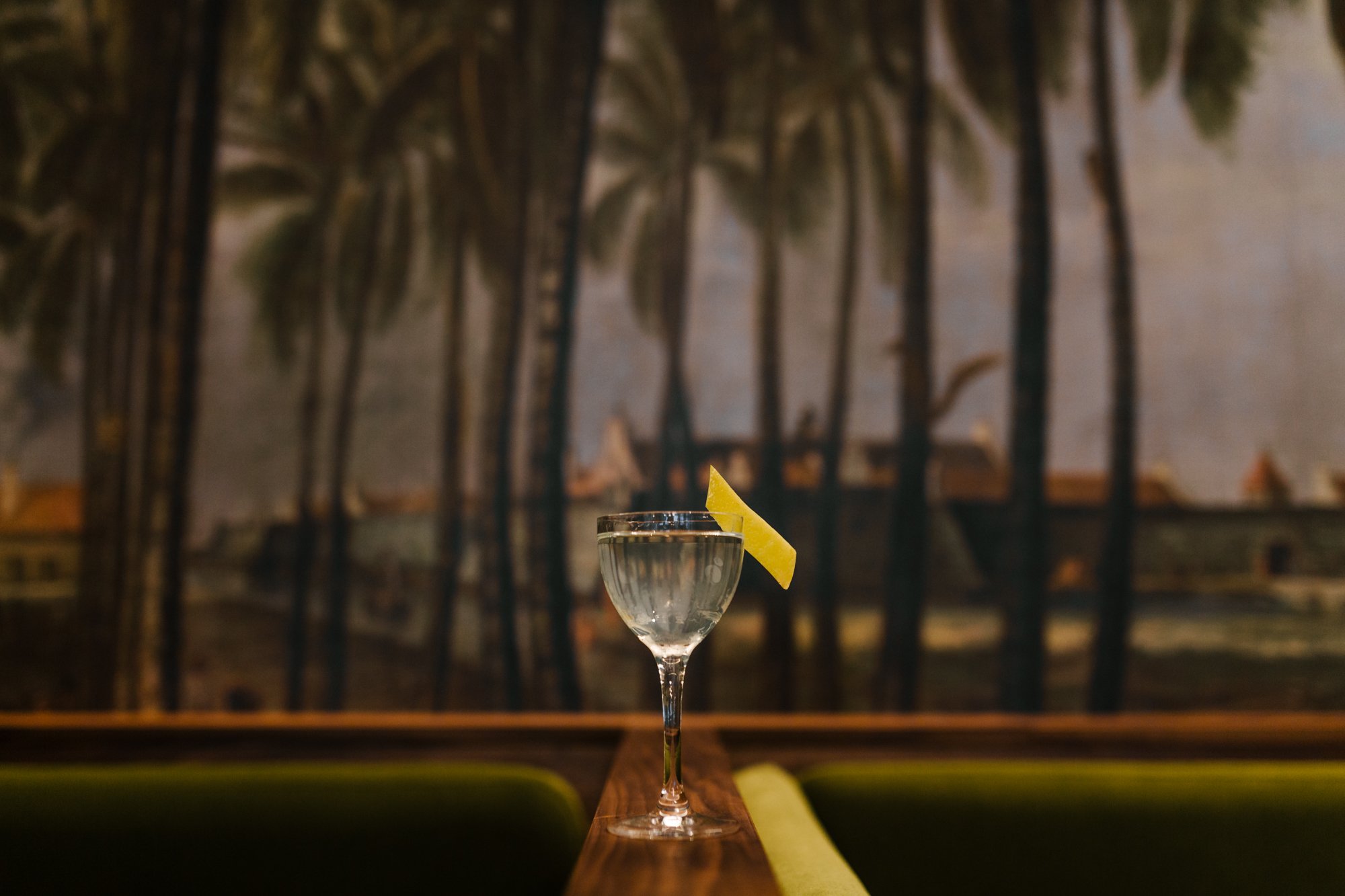 A cocktail glass with a lemon wedge garnish is placed on a wooden surface. In the background, there is a dimly lit outdoor setting with trees and a painted mural or backdrop.