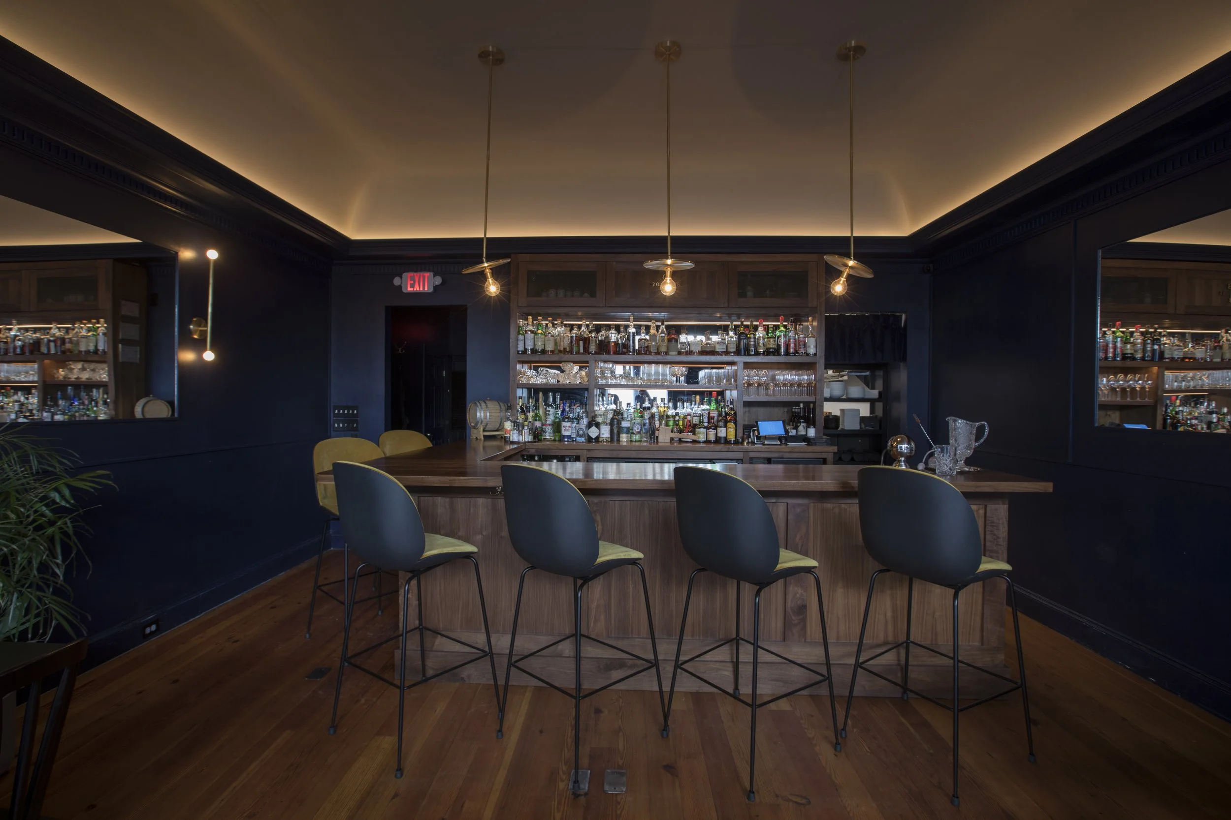Interior view of a bar with a wooden counter, filled with various liquor bottles, illuminated by hanging pendant lights. The walls are dark-colored with trays and glassware. There are chairs with dark legs and green cushions at the bar.