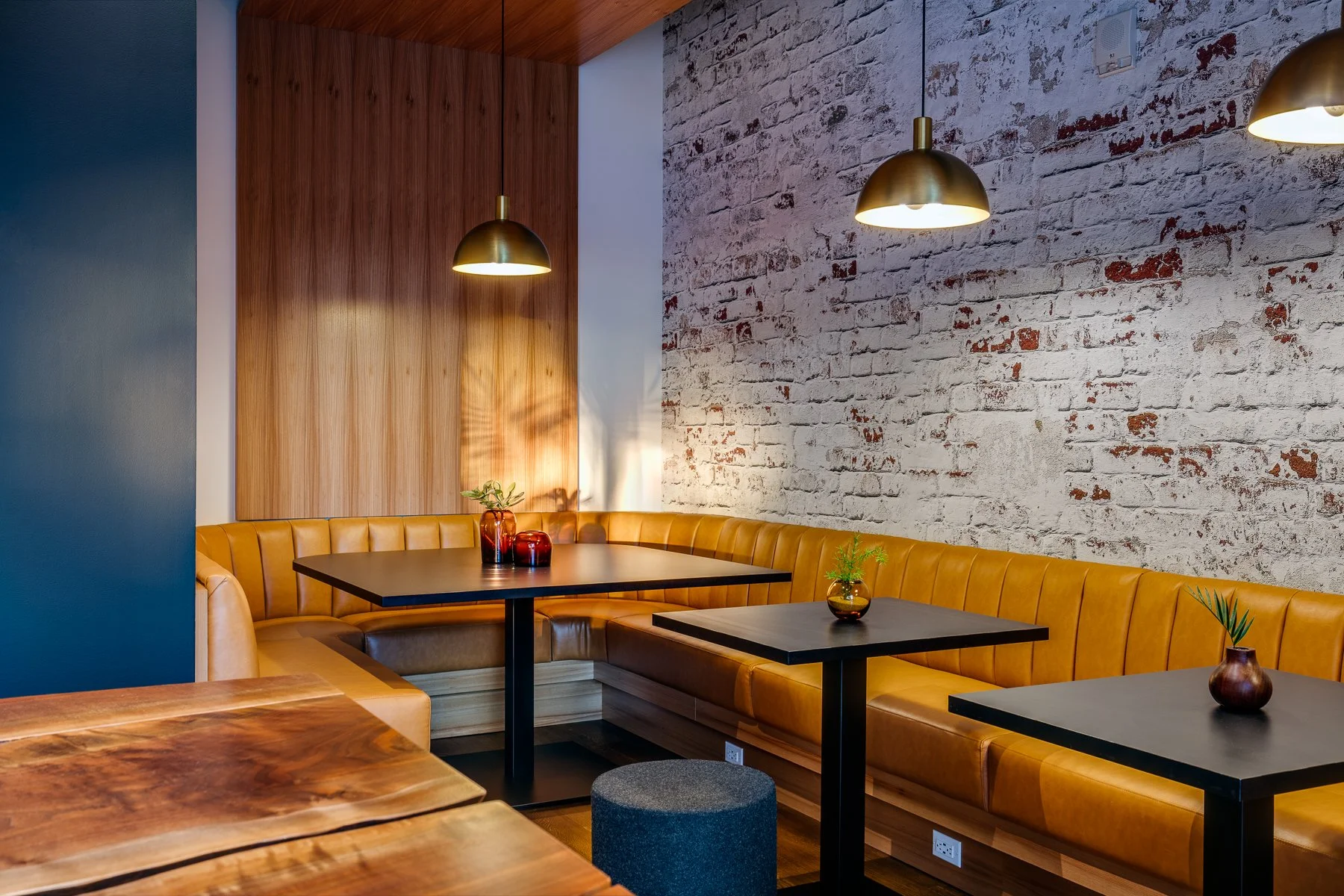 Interior of a restaurant with a corner booth, yellow leather seating, black tables, and decorative vases with plants. The wall features exposed white brick and wooden paneling, with pendant lights hanging overhead.