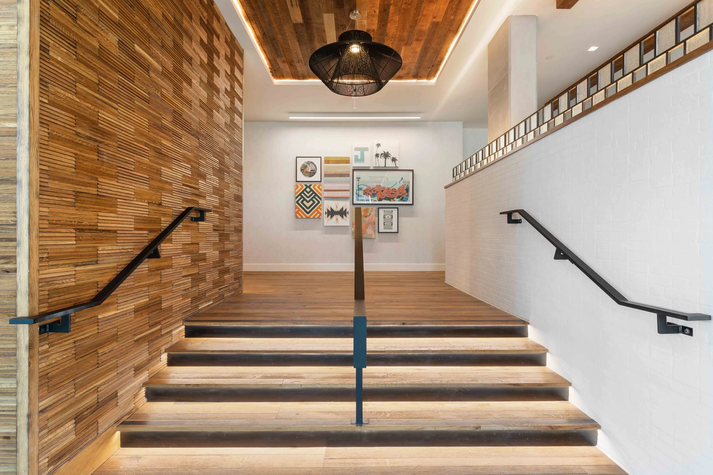 Interior staircase with black handrails, wooden steps, white brick wall on the right, wooden accent wall on the left, modern gallery wall with framed artworks, black pendant light, wooden ceiling with recessed lighting.