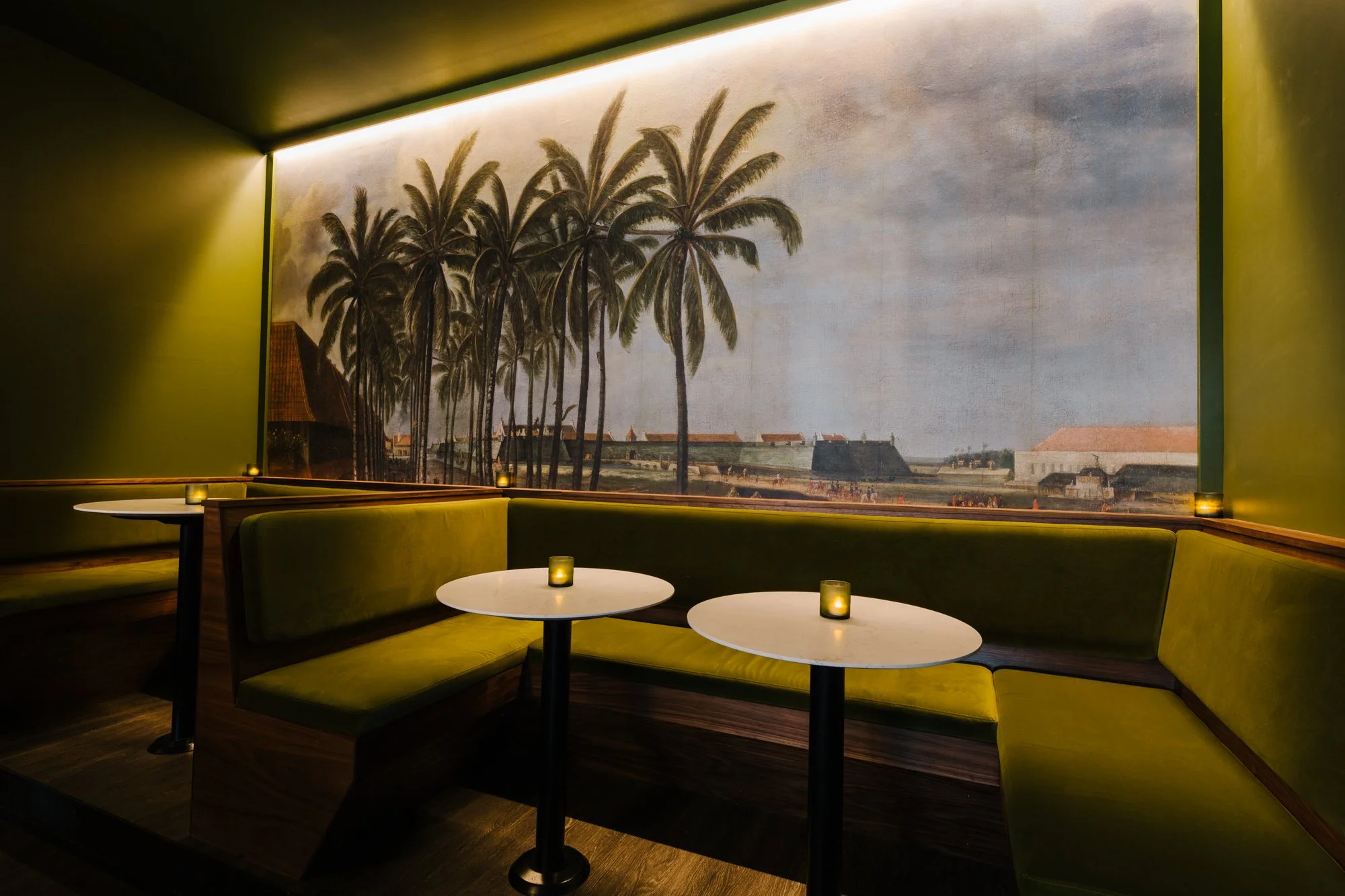 Indoor seating area with curved green banquettes, white round tables, candles, and a large mural of palm trees and a seaside village on the wall.