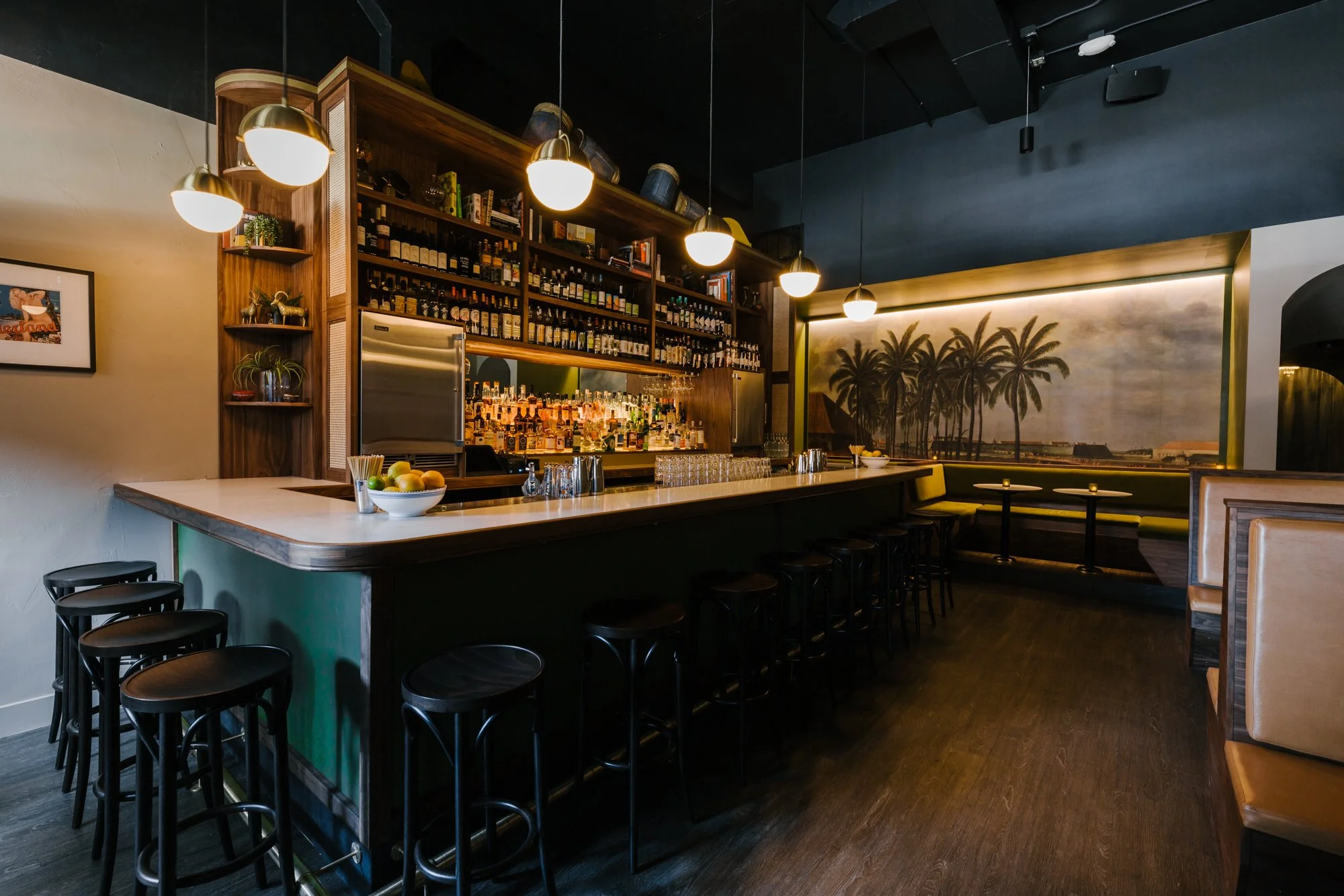 Interior of a modern bar with a high counter, black bar stools, and wooden shelves stocked with liquor bottles. Pendant lights hang from a black ceiling, and there's a large mural of palm trees on the wall behind the bar.