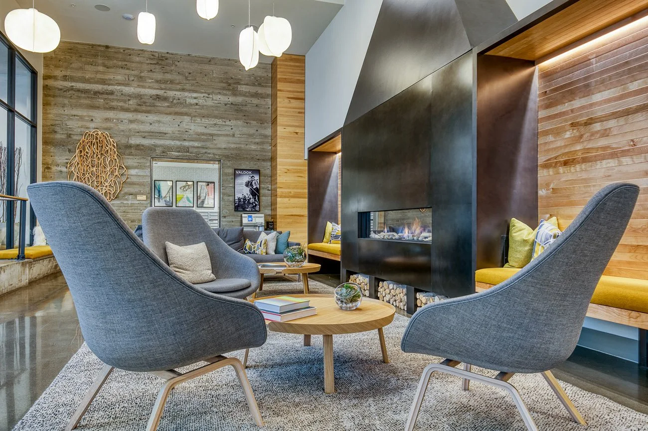 A modern lounge area with two gray armchairs, a wooden coffee table, and a fireplace. The space features wood paneling and decorative lighting.