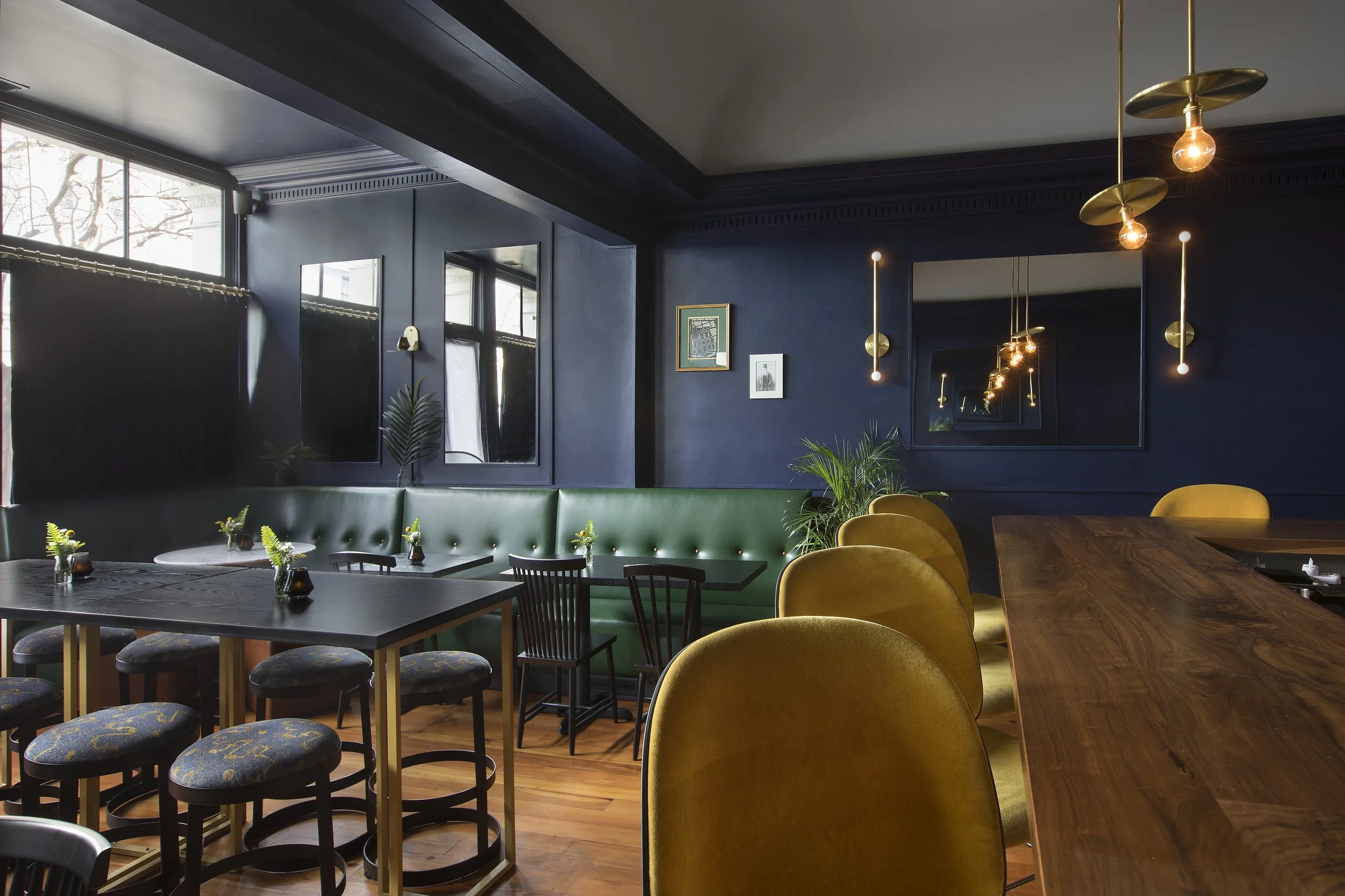 Interior of a modern restaurant or cafe with dark blue walls, hanging light fixtures, framed pictures, green plants, and various seating options including a green bench, wooden chairs, yellow velvet chairs, and barstools with patterned cushions.