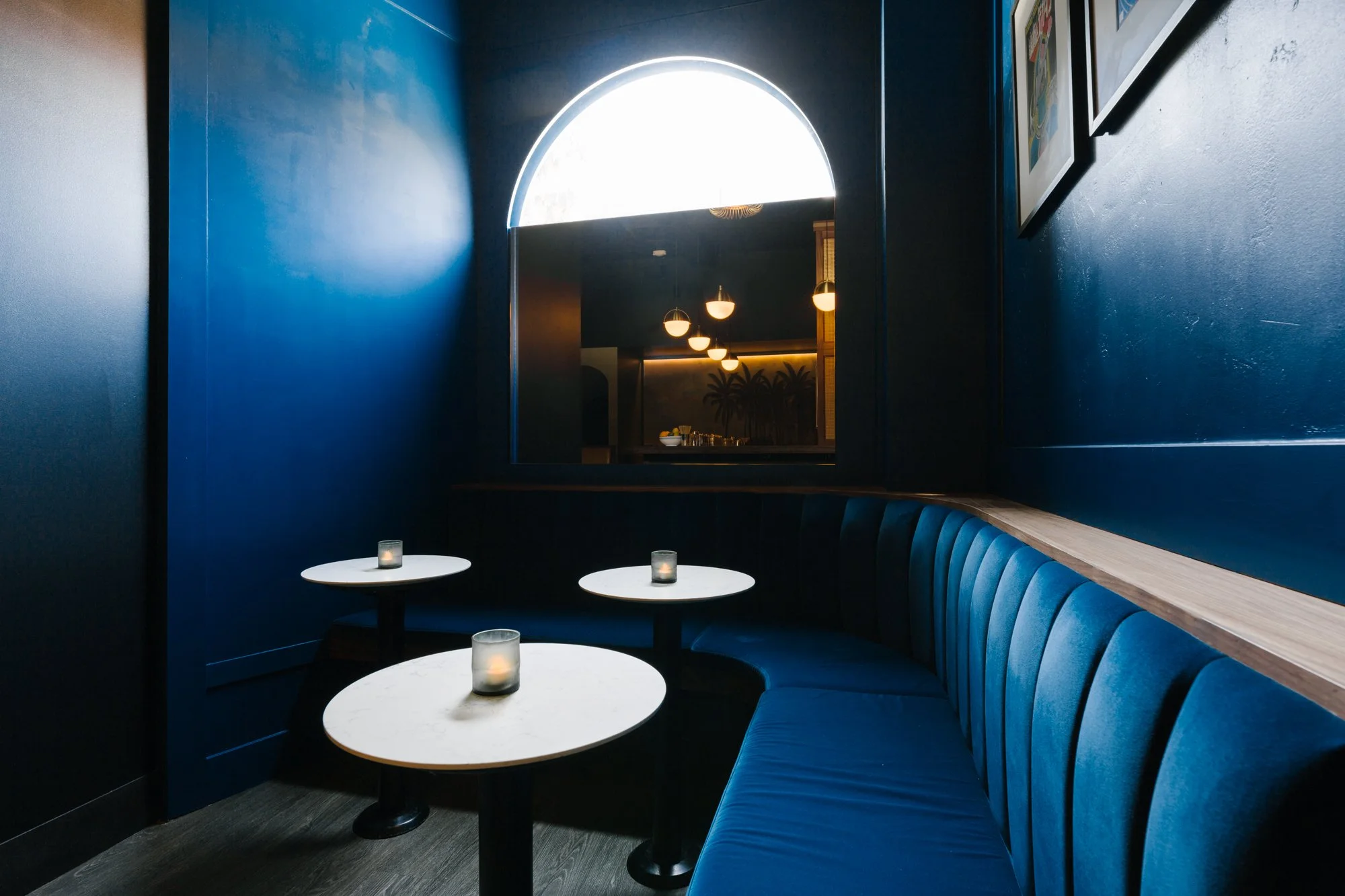 Interior of a modern restaurant with dark blue walls, a curved blue booth seating, and three white round tables each with a small candle holder. There is a large, semi-circular window above the entrance to another room that is illuminated by hanging pendant lights.