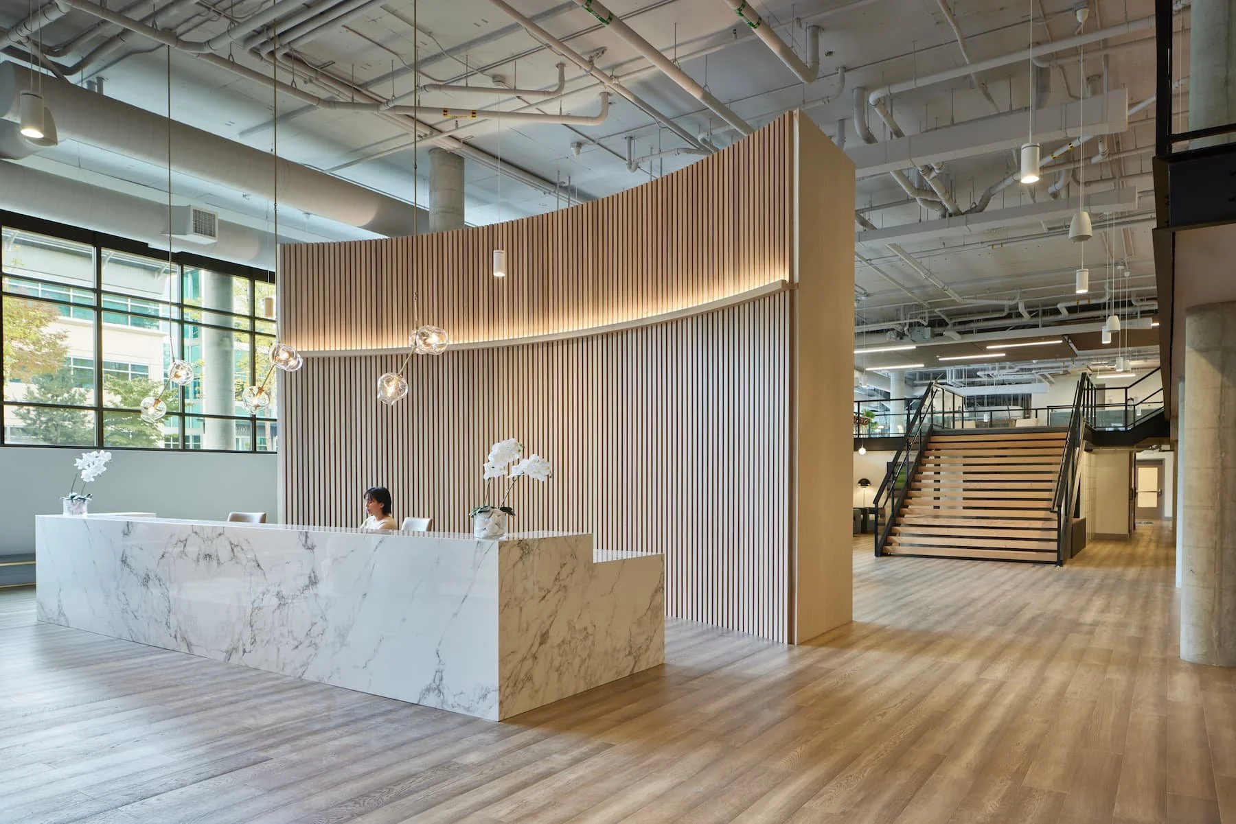 Modern office lobby with a marble reception desk, wood-paneled wall, pendant lighting, staircase with wooden treads and metal railing, large windows, and a woman sitting at the desk.