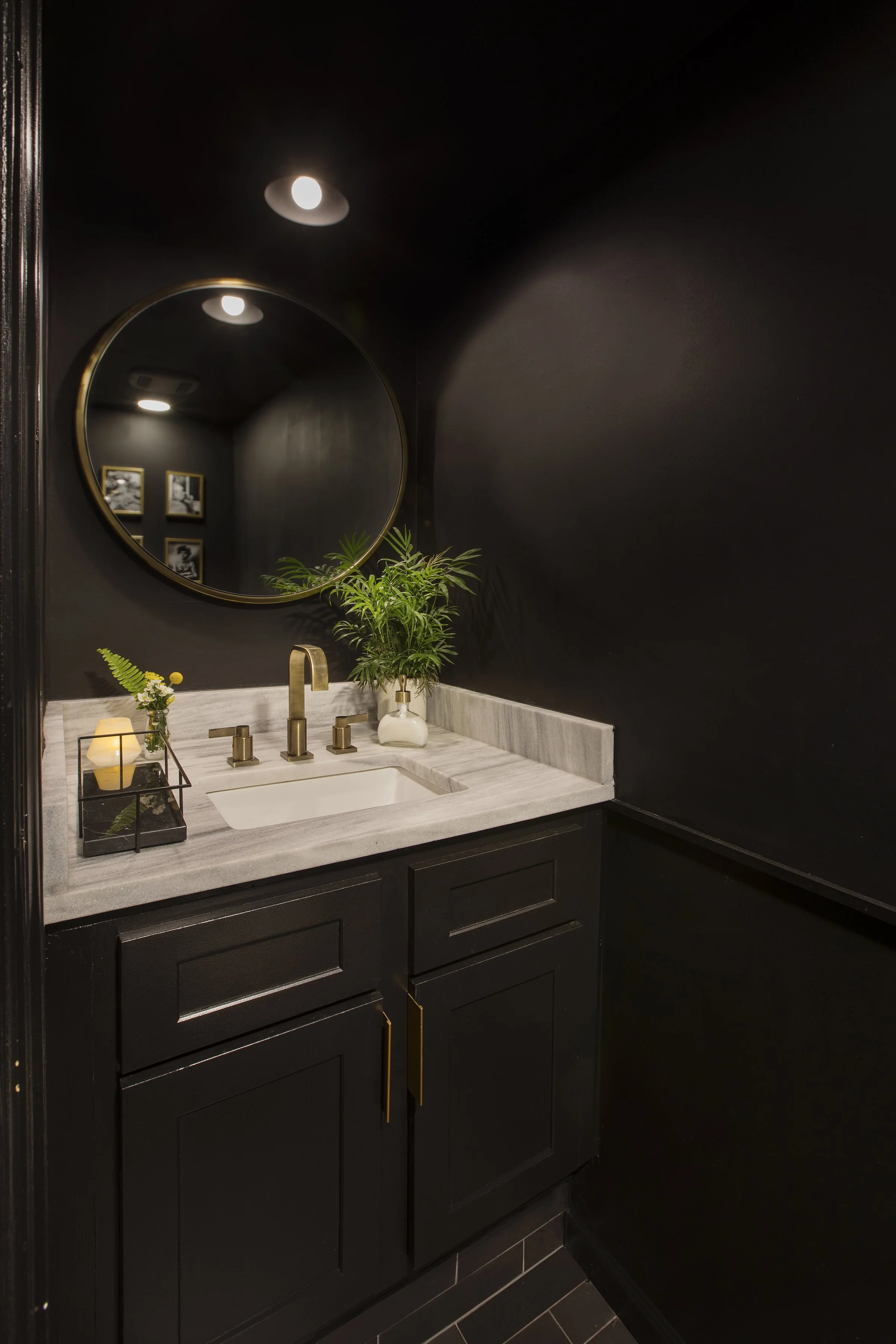 Black bathroom vanity with gold handles, marble countertop, and a round mirror. Decor includes a small candle, a tray with green fern, and a potted plant. Dark walls and framed black-and-white photos.