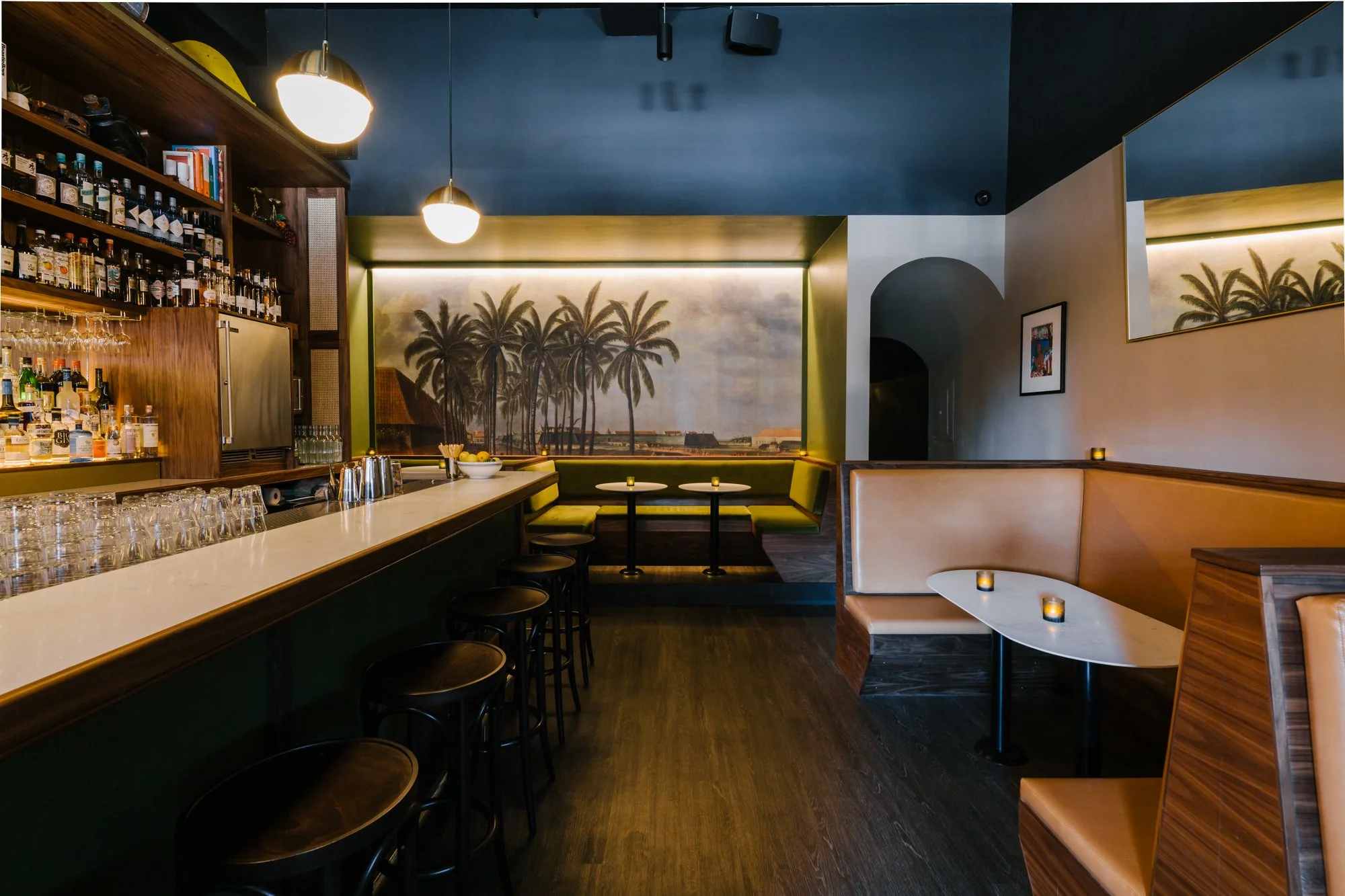 Interior of a modern bar or café with a long white countertop, shelves filled with bottles and glasses, a mural of palm trees on the wall, and cozy seating with small tables and candles.