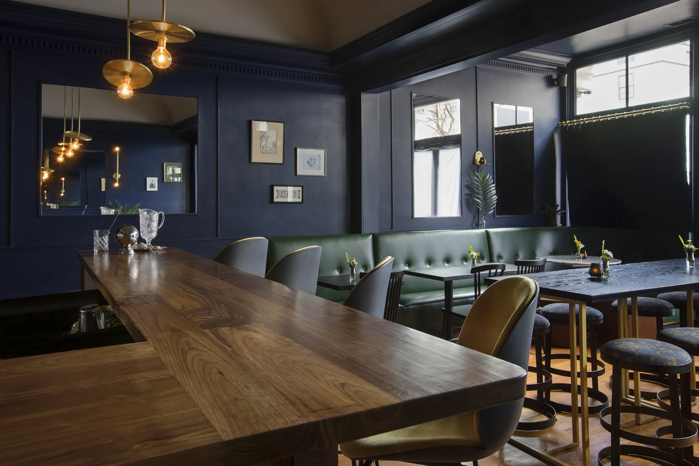 Interior of a modern restaurant with dark blue walls, large windows, and a blend of wooden and upholstered furniture, including a large wooden table and various chairs. There are framed pictures and a mirror on the wall, pendant lights hanging from the ceiling, and decorative plants.