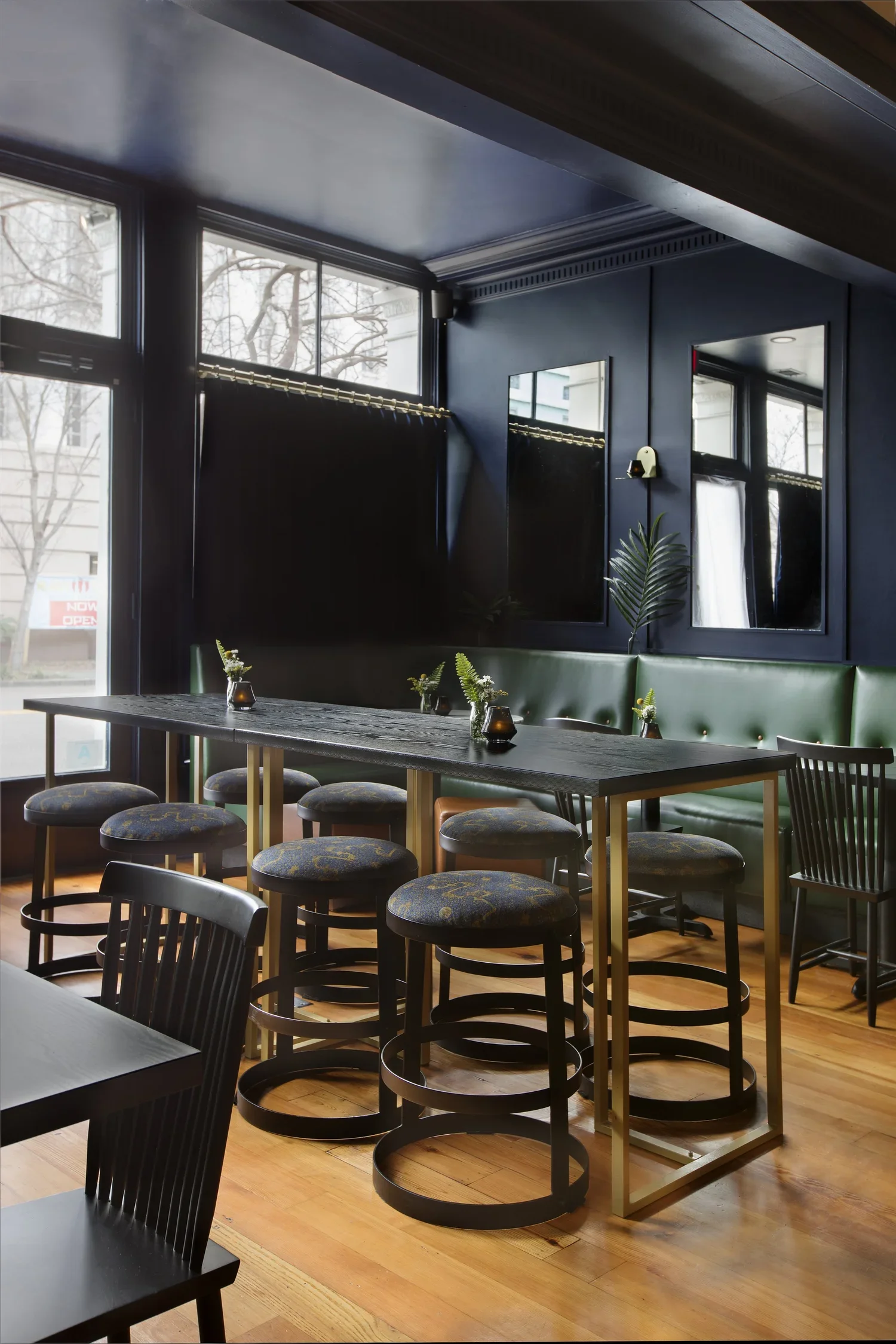 A modern restaurant interior with large windows, a wooden floor, a green upholstered bench, black chairs, a long table with small vases, and dark blue walls with mirrors and plants.