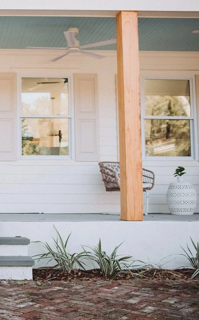 The image depicts a porch with a ceiling fan, a wooden support post, two windows, a wicker chair, a white decorative planter with a plant, and a small garden bed with ornamental grass in front of the porch. There are steps visible on the left side leading up to the porch. The house's exterior is painted white.