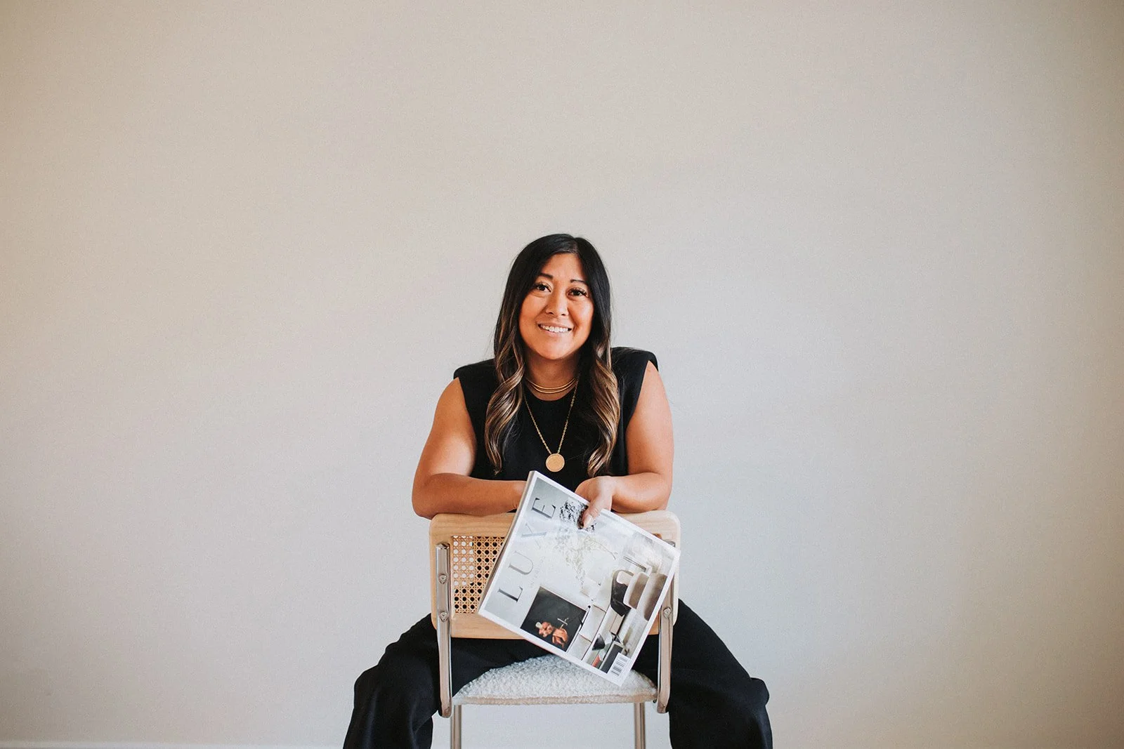 A woman sitting on a chair against a plain white wall, holding a magazine with the word 'Luxe' on the cover.