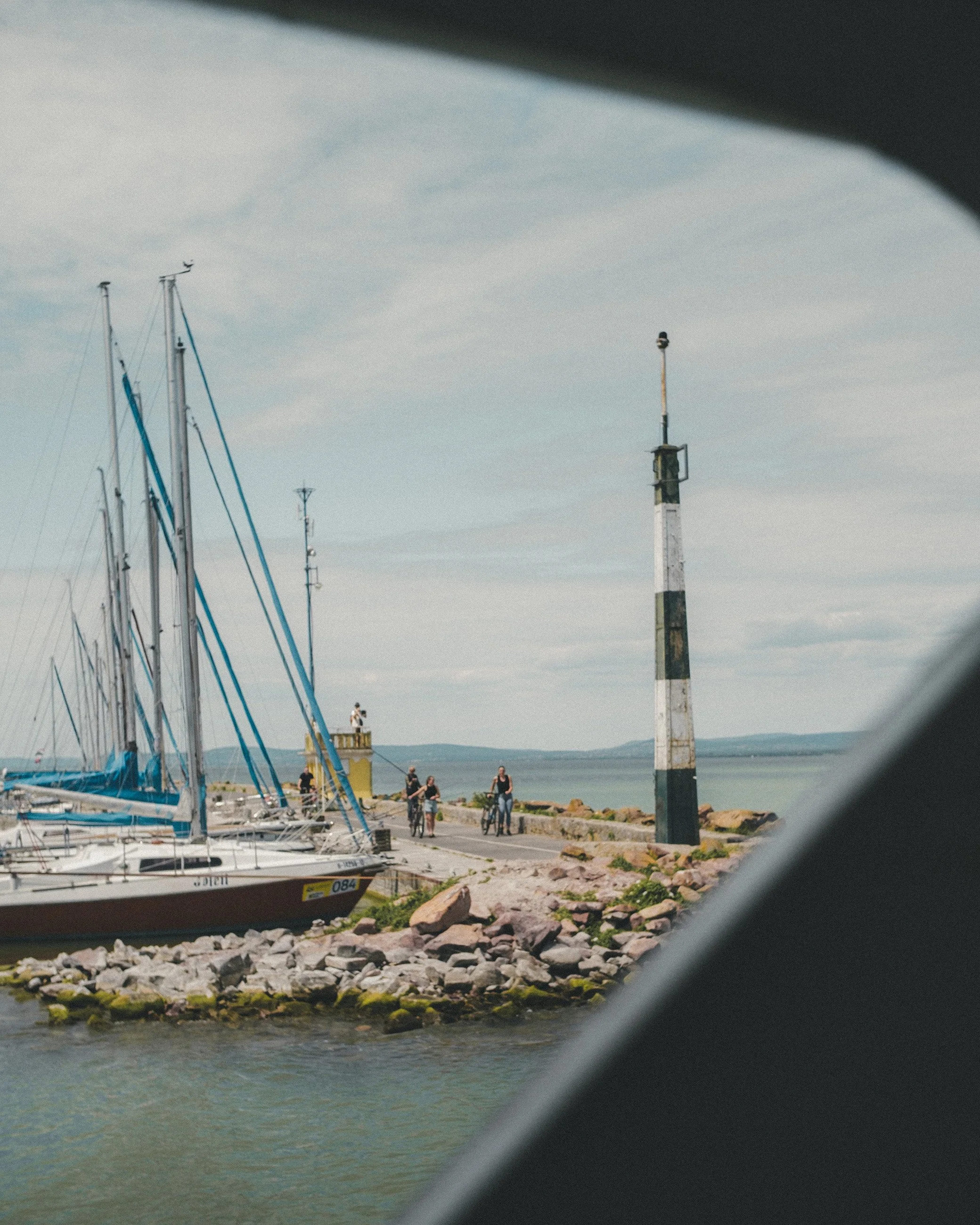 View of sailboats docked at a marina, with a rocky shoreline, a striped lighthouse, and a few people walking or riding bikes by the water, seen through a window or frame.