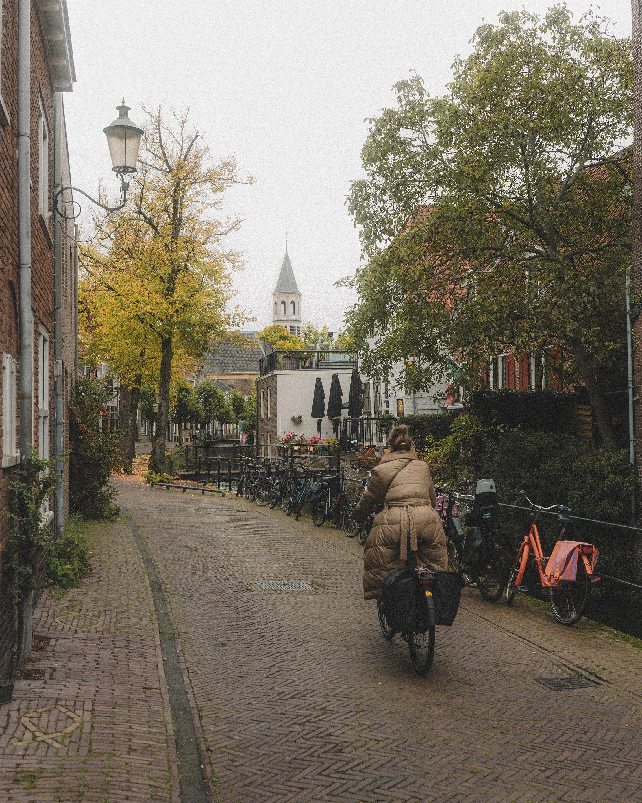 A woman riding a bicycle down a cobblestone street surrounded by parked bikes, trees with autumn leaves, and buildings with a church steeple in the background.