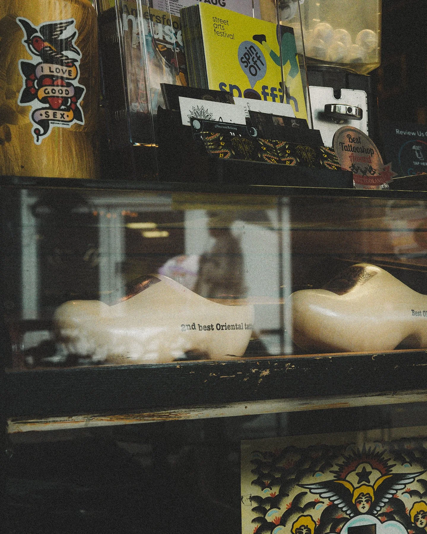 Display case with two tattoo foot molds labeled '2nd best Oriental tattoo' and various stickers, flyers, and artwork on top and behind the case.