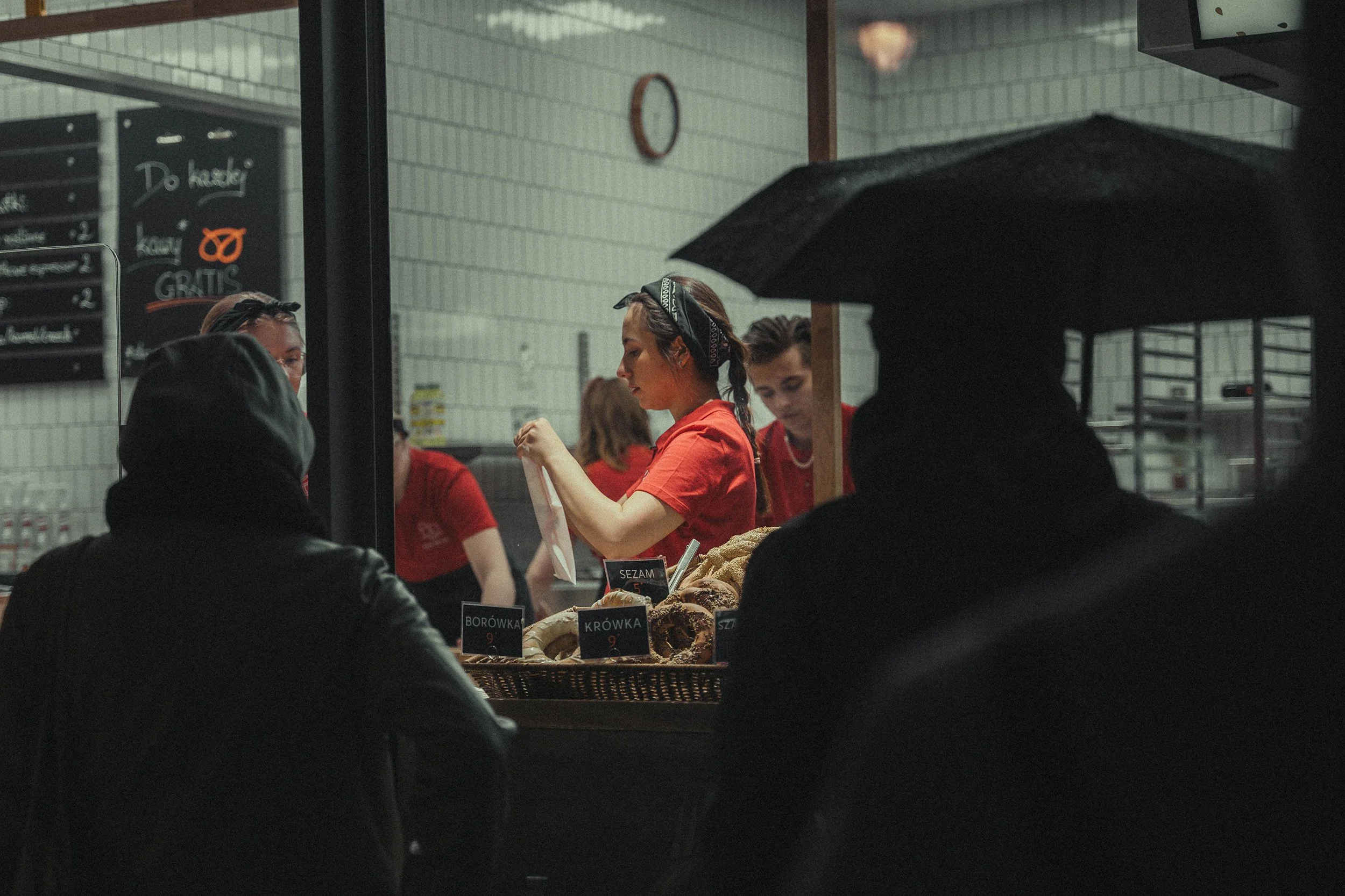 People waiting outside a bakery to order, with baked goods on display, viewed through a window, in a city setting.