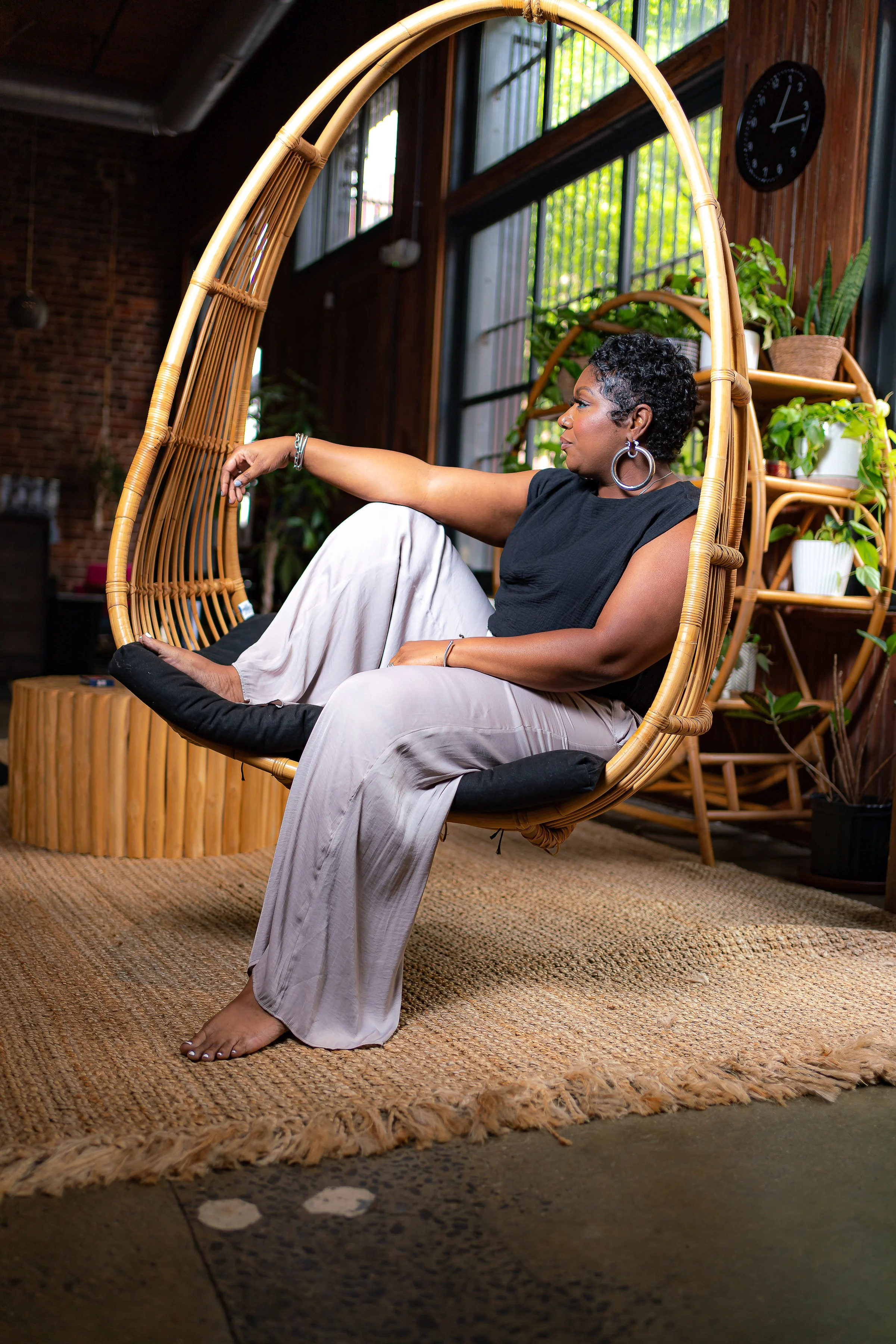 A woman with short curly hair sits relaxed in a hanging wicker chair with a black padded cushion, in a room with large windows, plants, a wooden clock, and brick walls, during daytime.