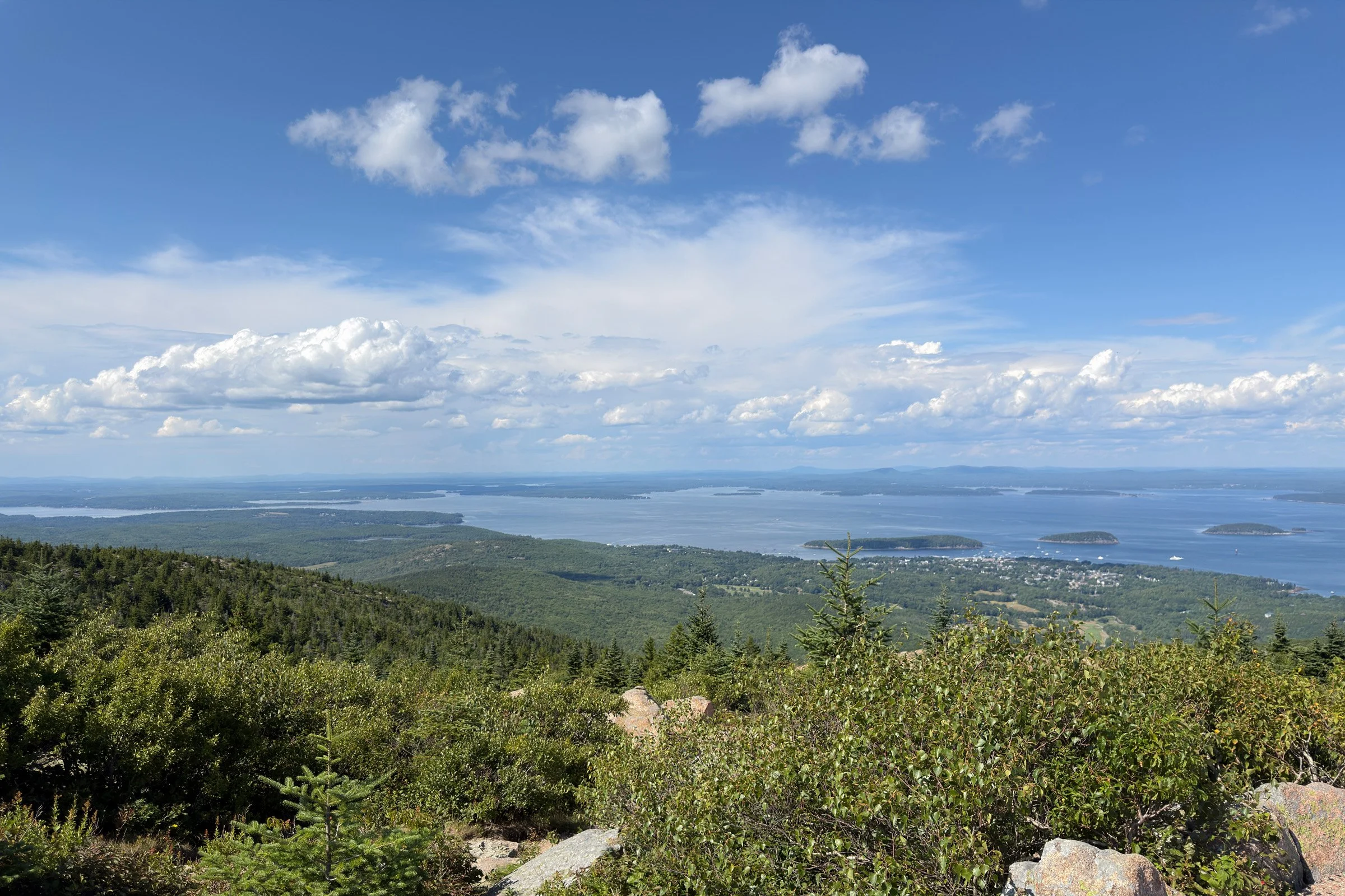 Islands in the Distance — Forested hills descend toward a blue bay dotted with islands beneath broad white clouds.