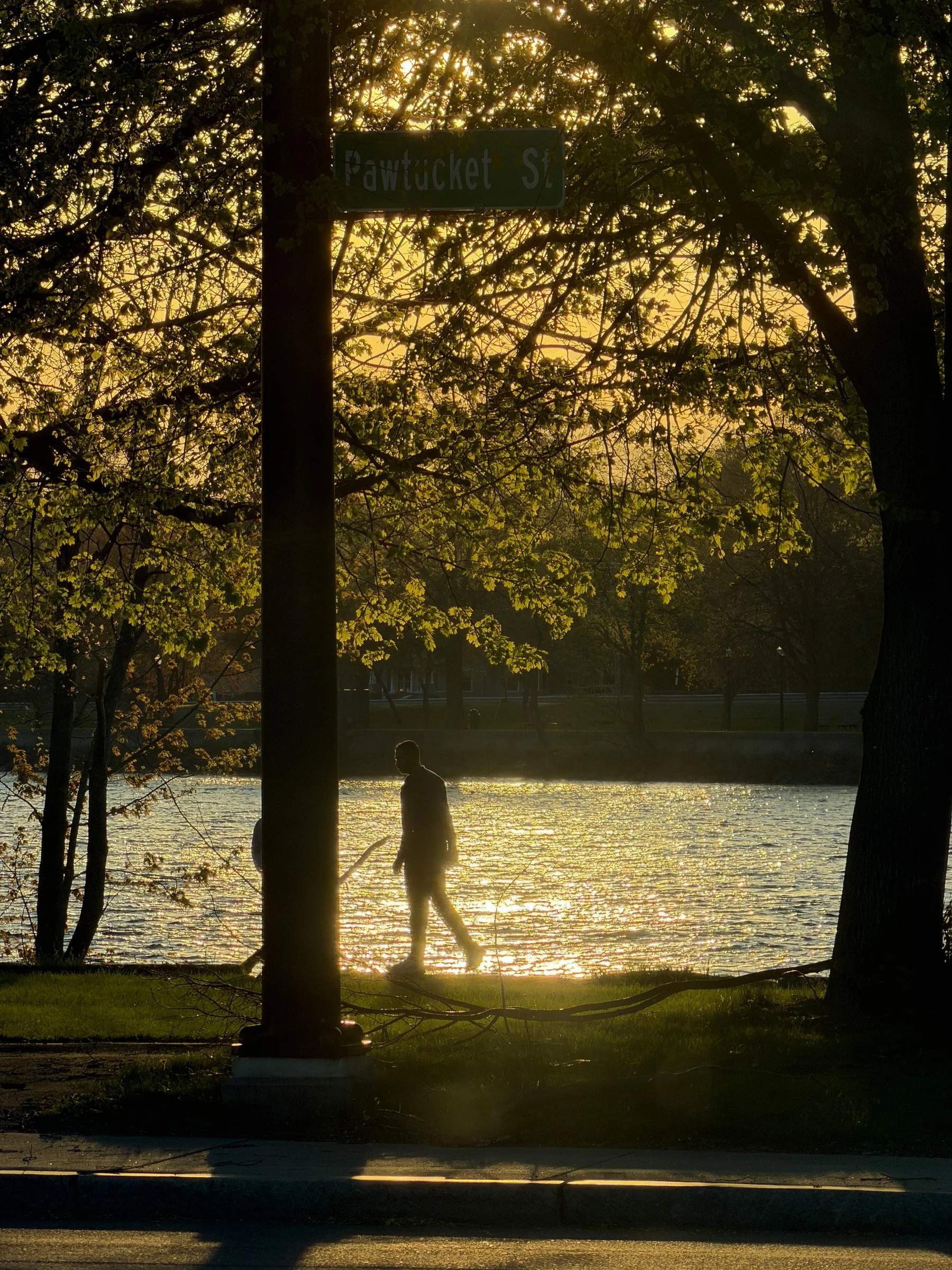Riverside Gold — A walking silhouette passes a sunlit river beneath leafy branches and a Pawtucket Street sign.