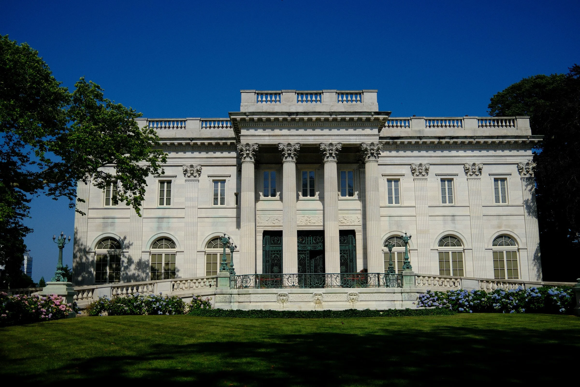 Summer Facade — A grand pale stone mansion with classical columns stands behind a lawn under a clear blue sky.