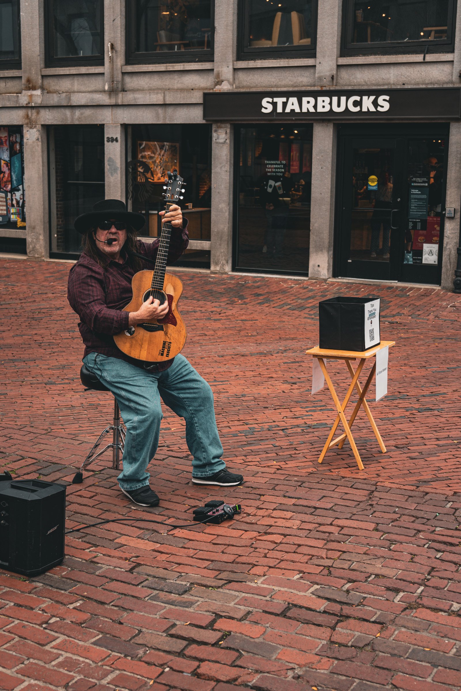 Street Melody — A guitarist in a black hat performs on a brick-paved street outside a cafe.