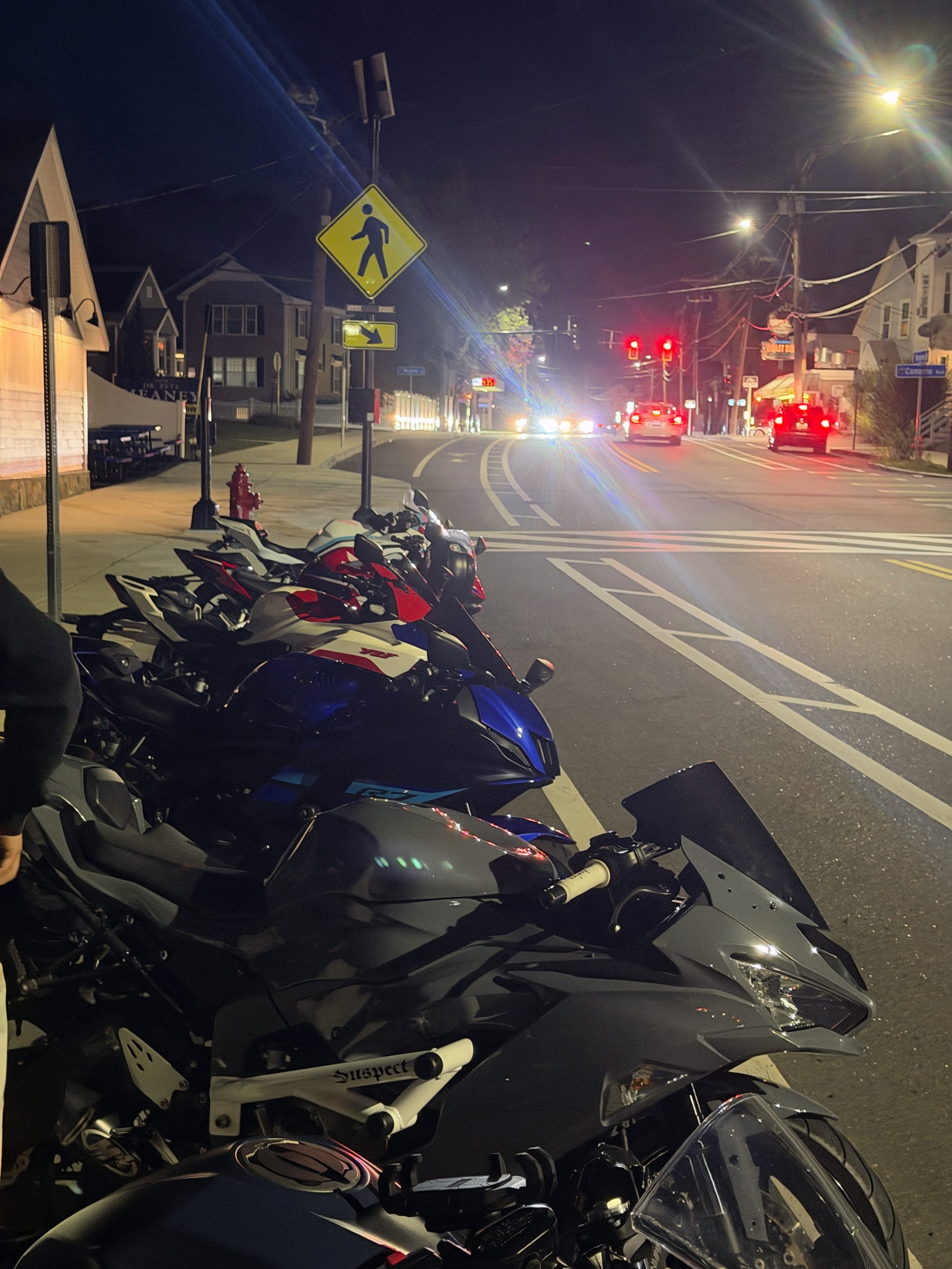Night Ride Lineup — A row of sport motorcycles lines a town sidewalk at night, with streetlights and traffic glowing beyond.