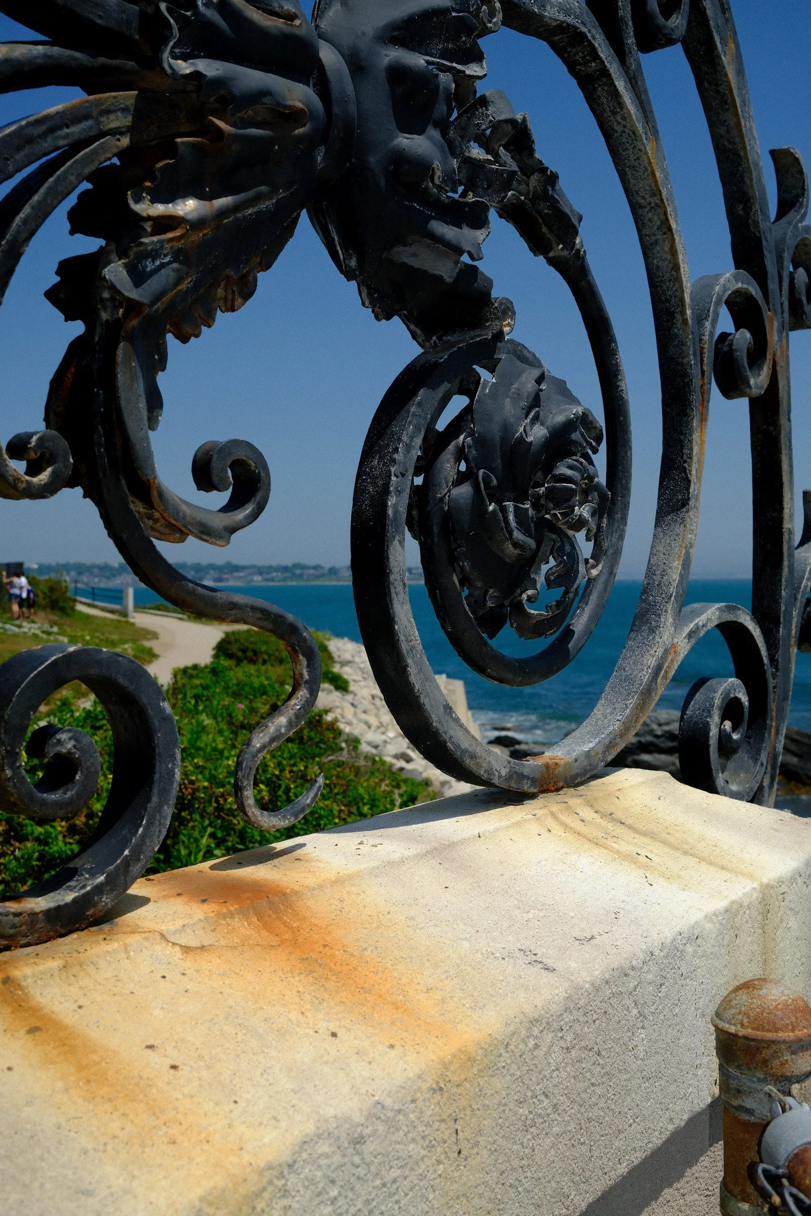 Iron Scrolls — Ornate black ironwork casts sculptural curves against pale stone and a blue coastal sky.