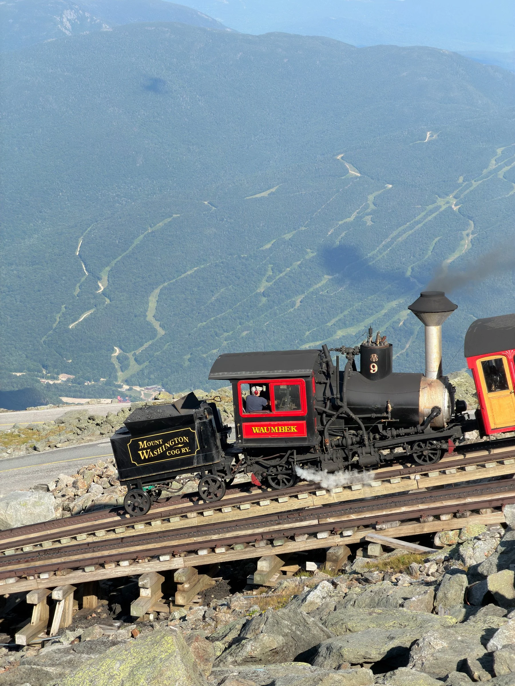 Summit Steam — A black steam locomotive with a red cab sits on steep mountain tracks above a hazy green valley.