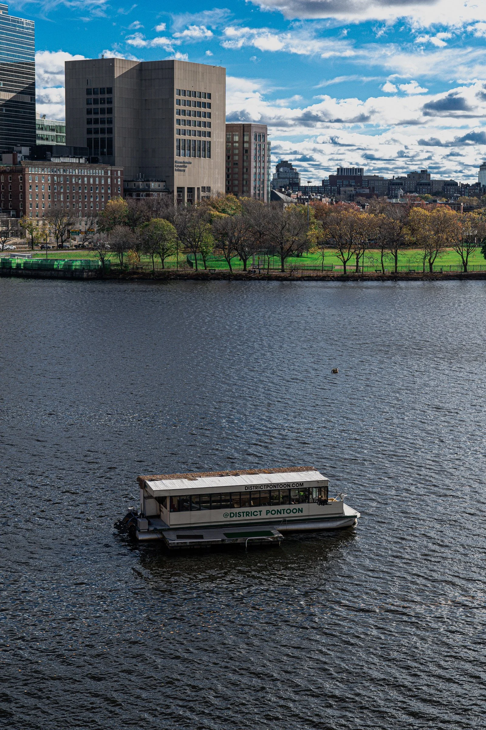 Across the River — A small pontoon boat crosses the river below autumn trees and a Boston skyline.