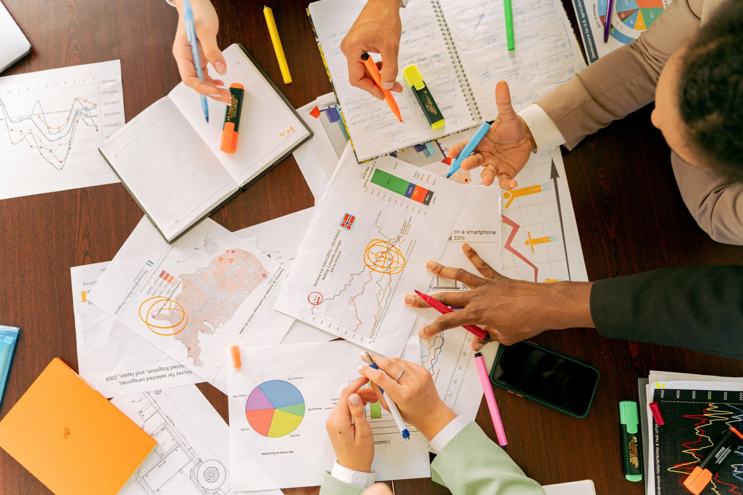 People working on a desk with charts, graphs, notebooks, highlighters, and pens, collaborating on data analysis and planning.