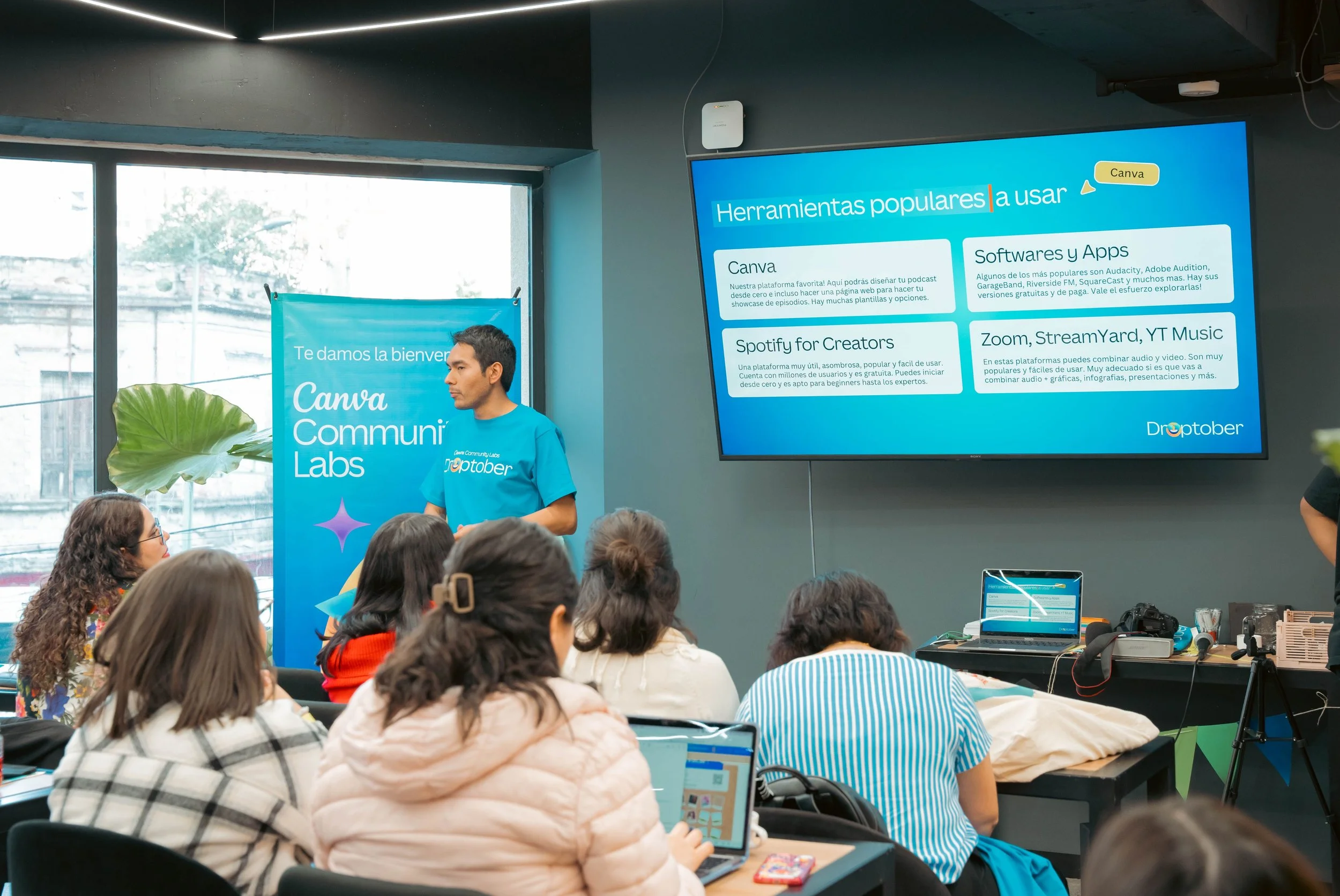 A group of people attending a presentation or workshop with large screens displaying information about popular tools like Canva, Spotify for Creators, and Zoom, StreamYard, YT Music. The setting appears to be a conference room or classroom with a presenter standing near the screens.