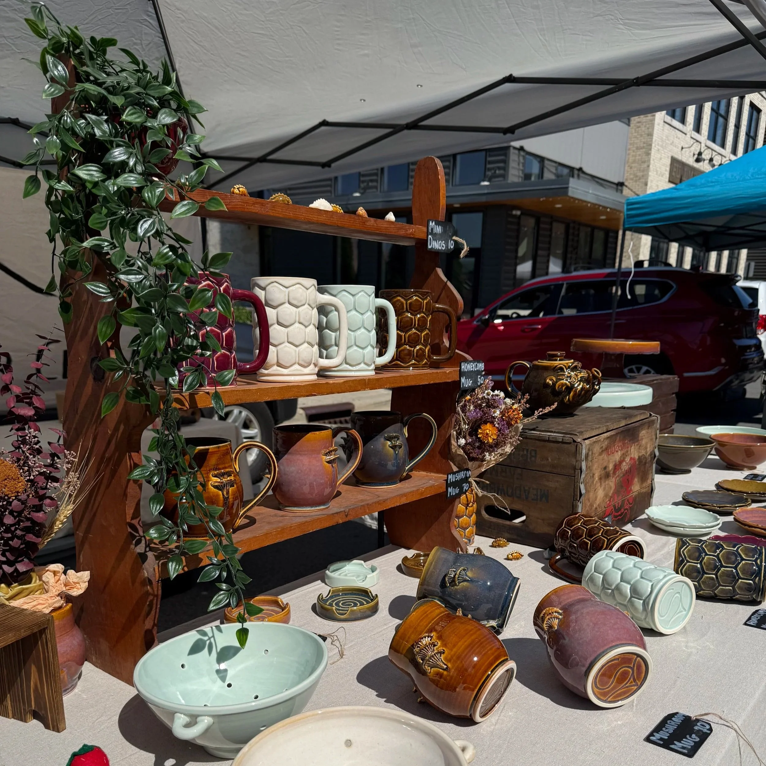 Outdoor market stall displaying ceramic mugs, bowls, and vases with a potted plant on the left, under a canopy, in an urban setting with buildings and a red vehicle in the background.