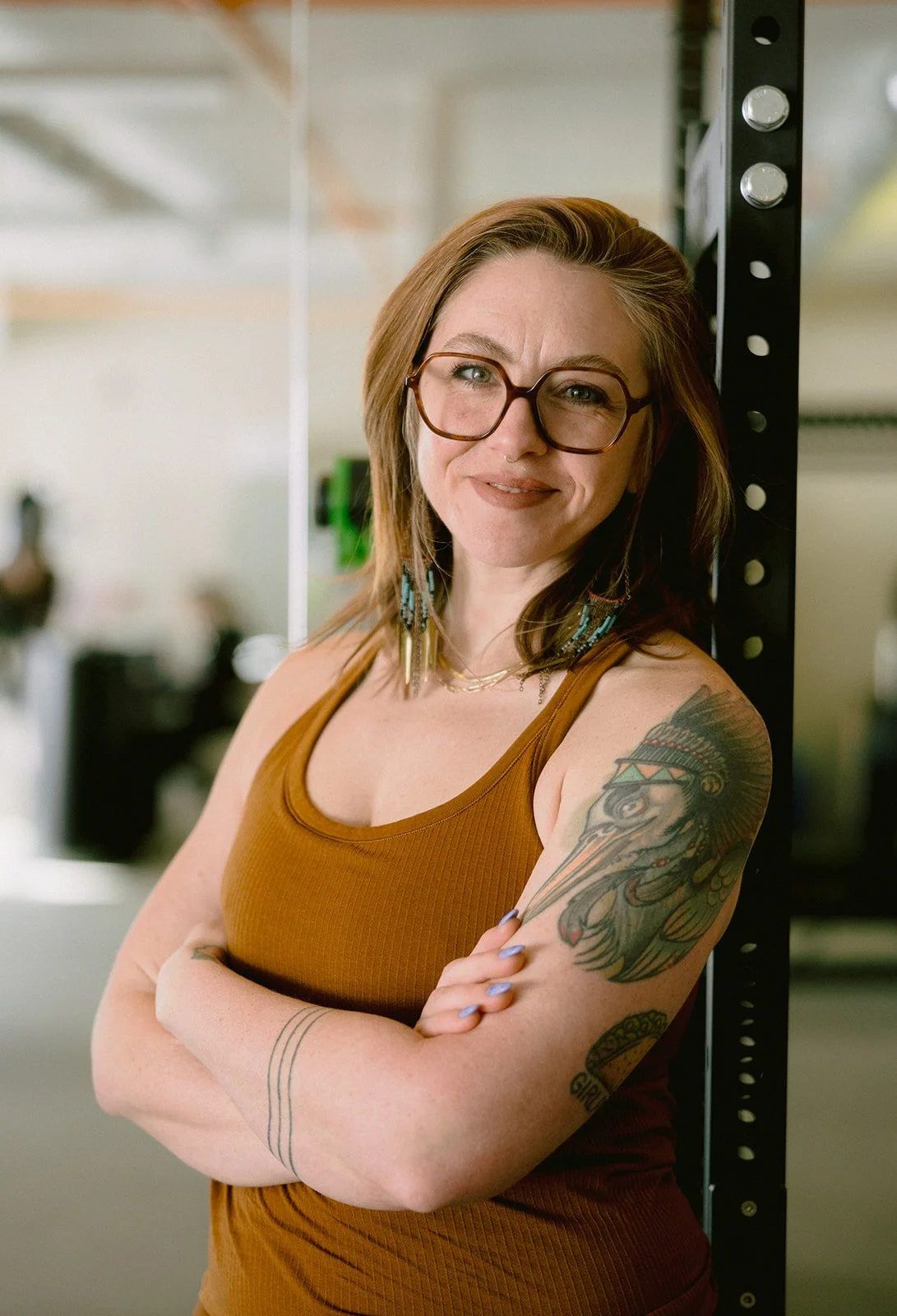 Smiling woman with shoulder-length red hair, wearing large eyeglasses, gold jewelry, and a black sleeveless top, with a tattoo on her right arm, in a well-lit room.