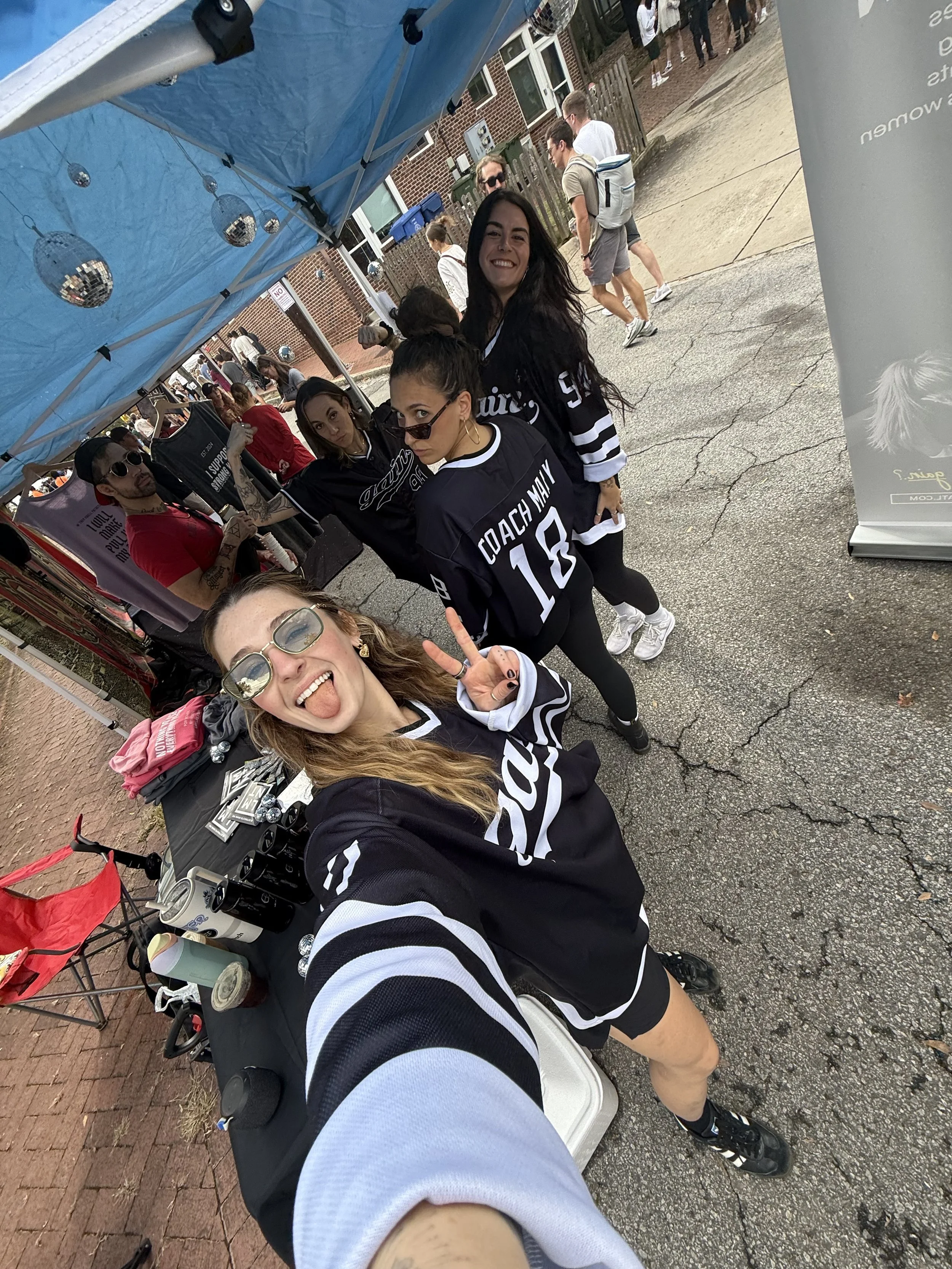 Group of friends taking a selfie outdoors at an event, with some wearing hockey jerseys, under a blue canopy with disco ball decorations.