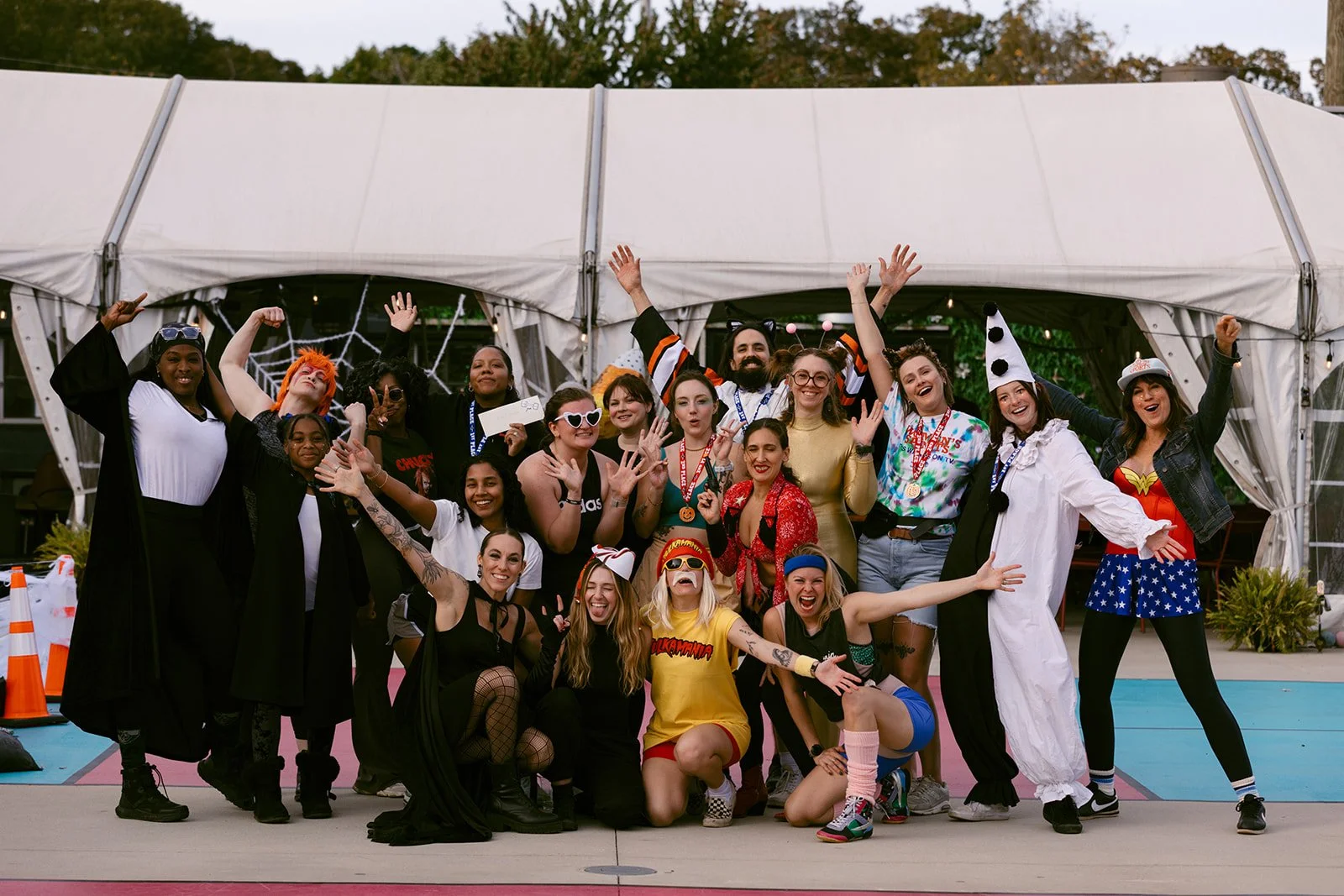 Group of people in costumes celebrating at a costume party outdoor, some with playful accessories, standing in front of a white tent.