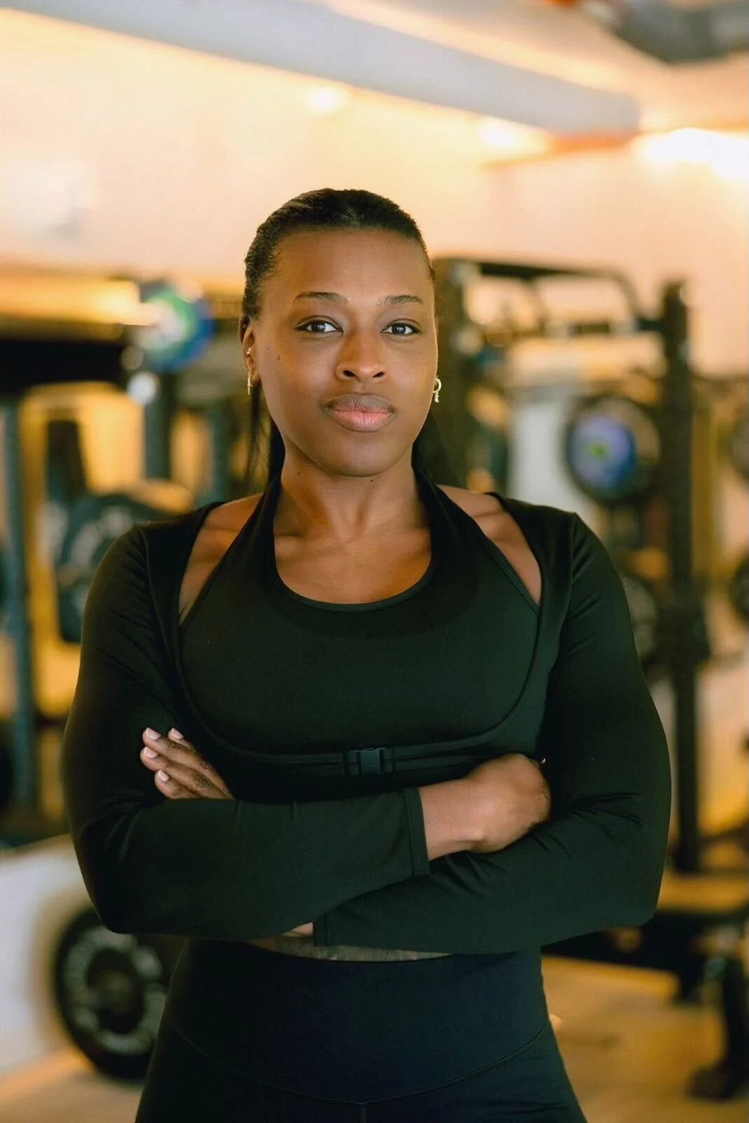 A confident woman with dark skin and black hair tied back, wearing a black athletic jacket and sports bra, standing with arms crossed against a plain light background.