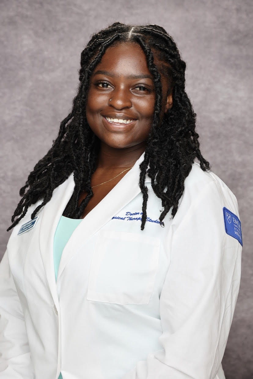 A smiling woman in a white medical coat with a name tag and a hospital logo, standing against a gray background.