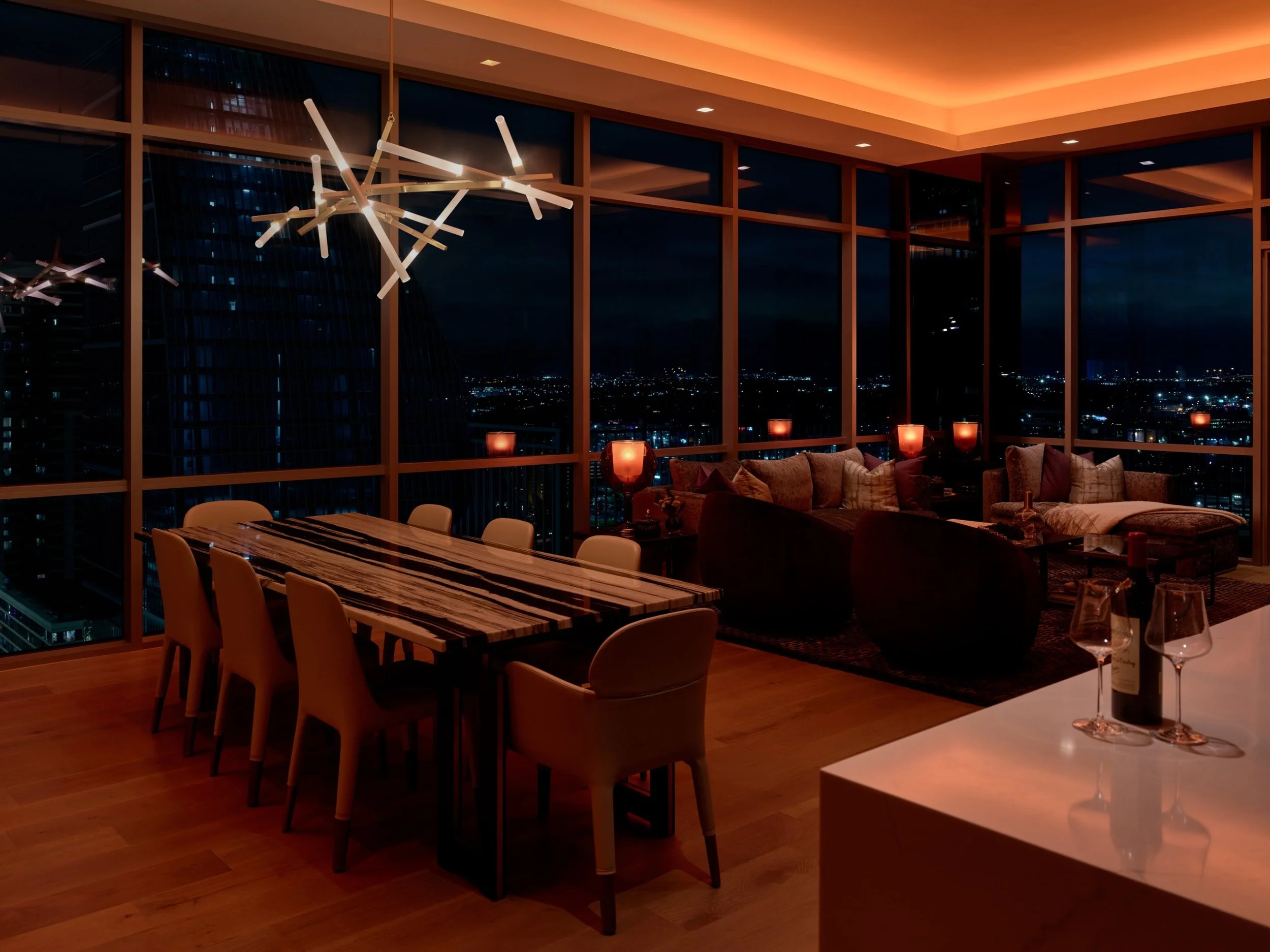 A luxury high-rise living and dining space featuring Lutron QS shades, HomeWorks QSX control, Lumaris linear lighting in the ceiling cove, and Ketra fixtures for layered, dynamic illumination. The space includes a modern dining table with seating for
