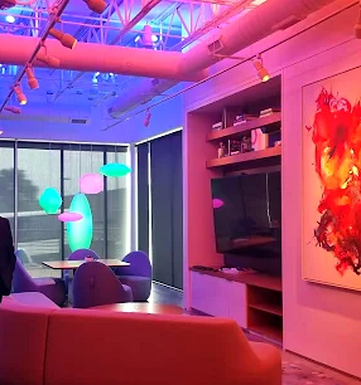Modern living room with multicolored neon lighting, purple chairs around a small dining table, large window with vertical blinds, floating light fixtures, and a wall-mounted flat-screen TV.