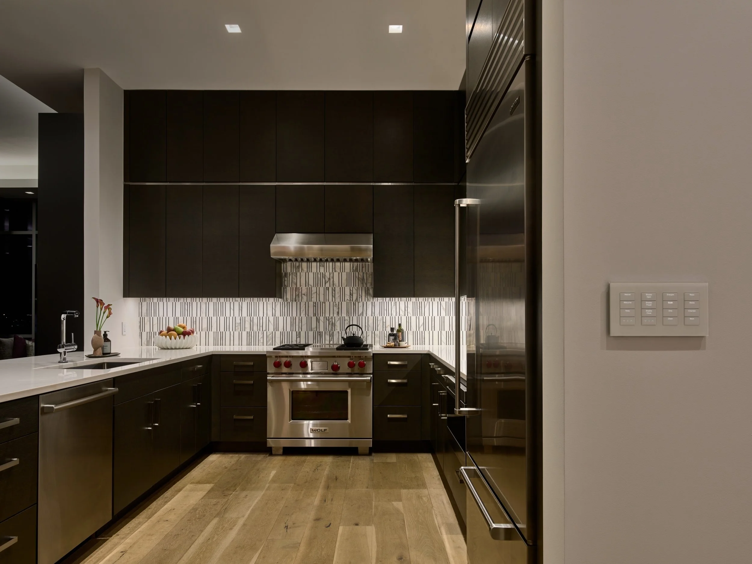 A sleek modern kitchen with Lutron smart lighting control balancing task and ambient illumination. The recessed LED downlights provide even overhead lighting, while the Lutron wall keypad near the entry allows instant scene control for cooking, enter