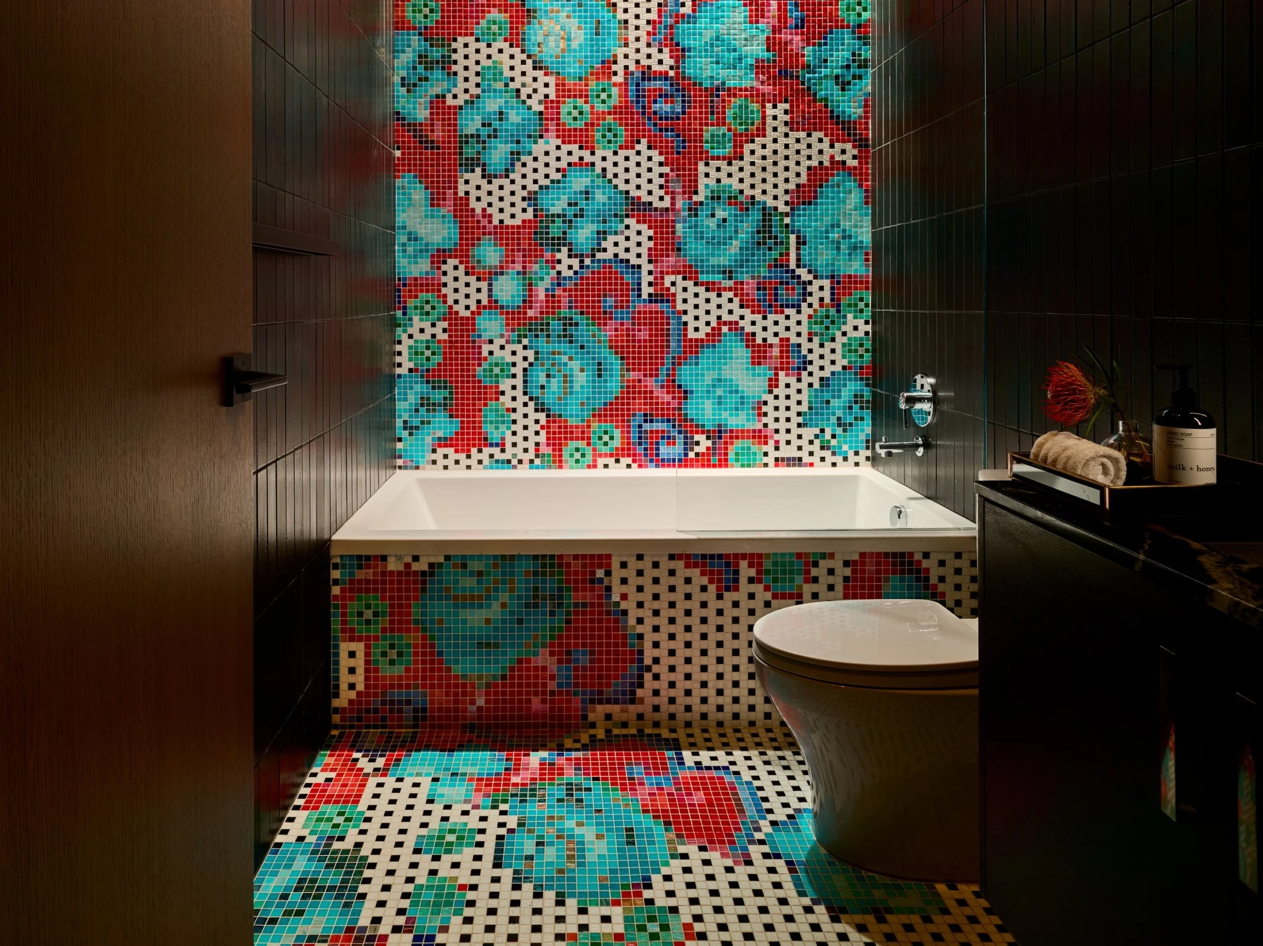 A vibrant bathroom showcasing Lutron smart lighting that enhances bold mosaic tilework in red, blue, and turquoise floral patterns across the floor and tub surround. Subtle recessed LED downlighting highlights the textures and colors, while dark mode