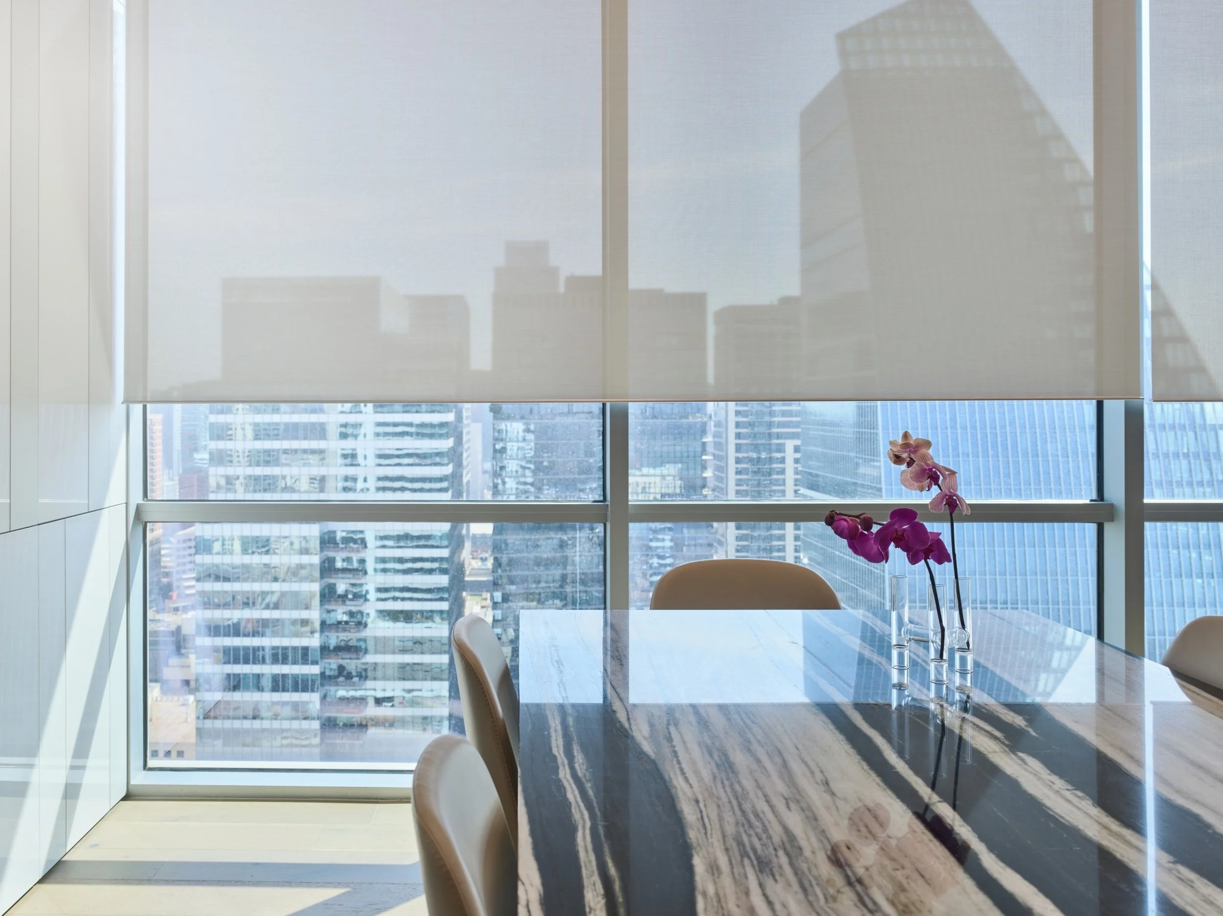 A modern high-rise dining or meeting space with Lutron motorized roller shades filtering daylight across floor-to-ceiling glass windows. The shades reduce glare while maintaining views of the surrounding city skyline, blending privacy with openness.