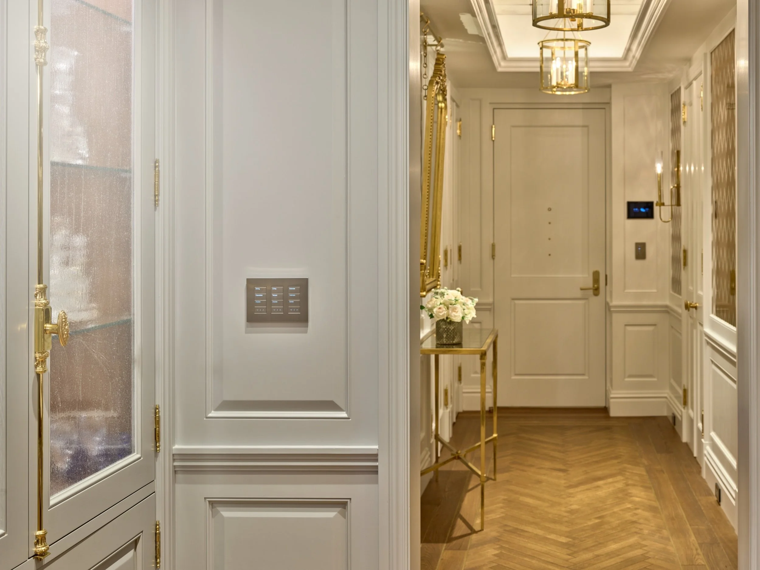 An elegant foyer illuminated by Lutron smart lighting control, blending classic design with modern convenience. The space features herringbone wood floors, crisp white paneled walls with gold hardware, and a console table with floral arrangement. A L