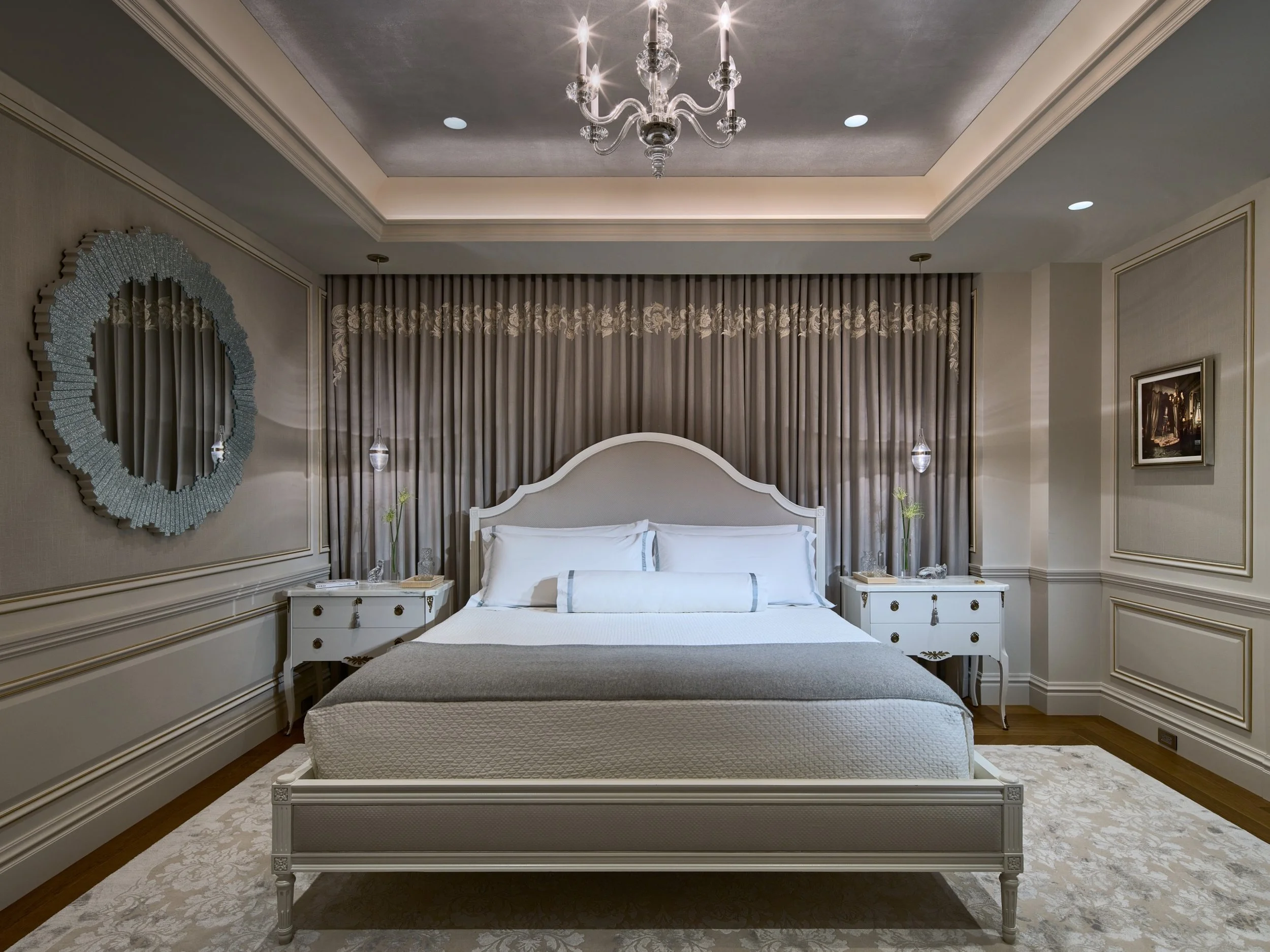 An elegant bedroom designed with layered Lutron lighting control to highlight architectural details and create a luxurious retreat. Recessed LED downlights softly illuminate the coffered ceiling, while a crystal chandelier adds sparkle and warmth. Wa