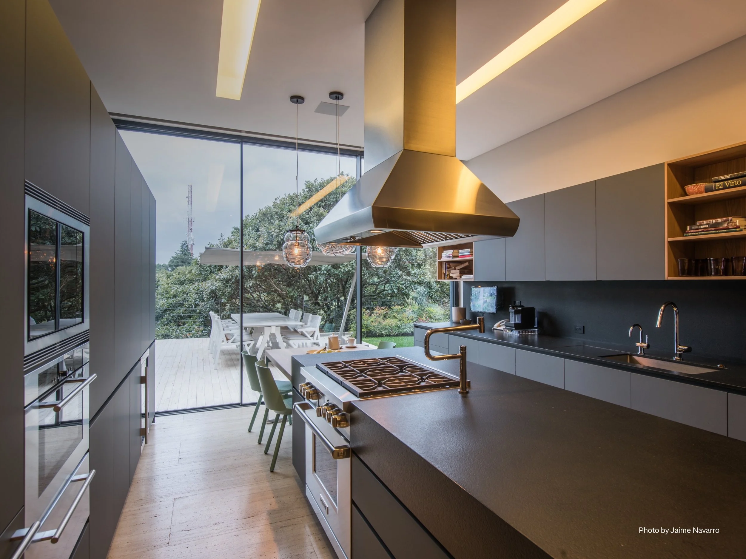 A sleek modern kitchen illuminated by Lutron smart lighting with a mix of linear recessed fixtures and pendant lights over the island. Expansive floor-to-ceiling glass doors open to a patio dining area, with the option for motorized shades to manage