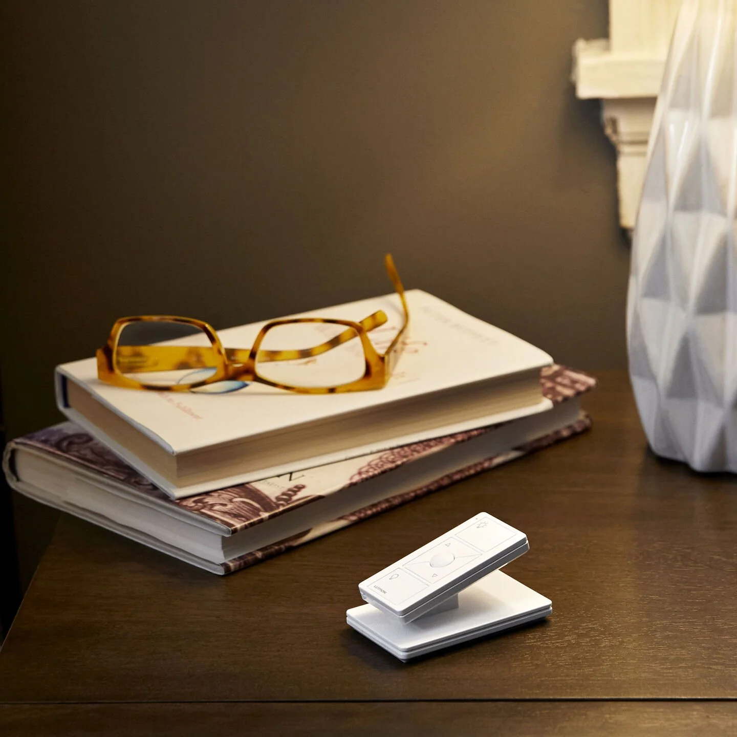 A pair of tortoiseshell glasses resting on top of a stack of three books on a wooden desk. To the right, there is a white geometric-patterned decorative object and a white remote control.
