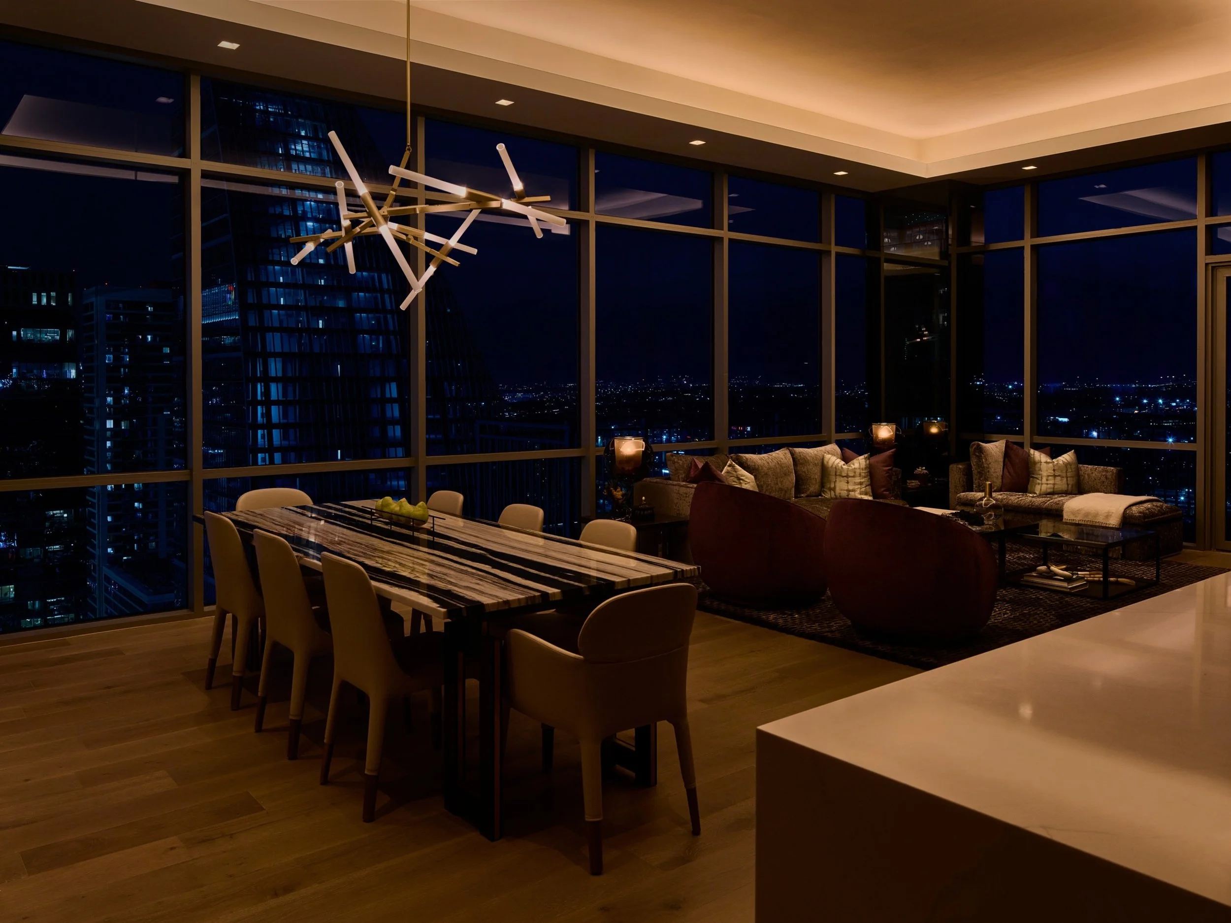 A high-rise dining and living space illuminated with Ketra D2 downlights and Lumaris tape lighting integrated into the ceiling cove, creating a layered nighttime scene. The room features a modern linear chandelier above the dining table, with expansi