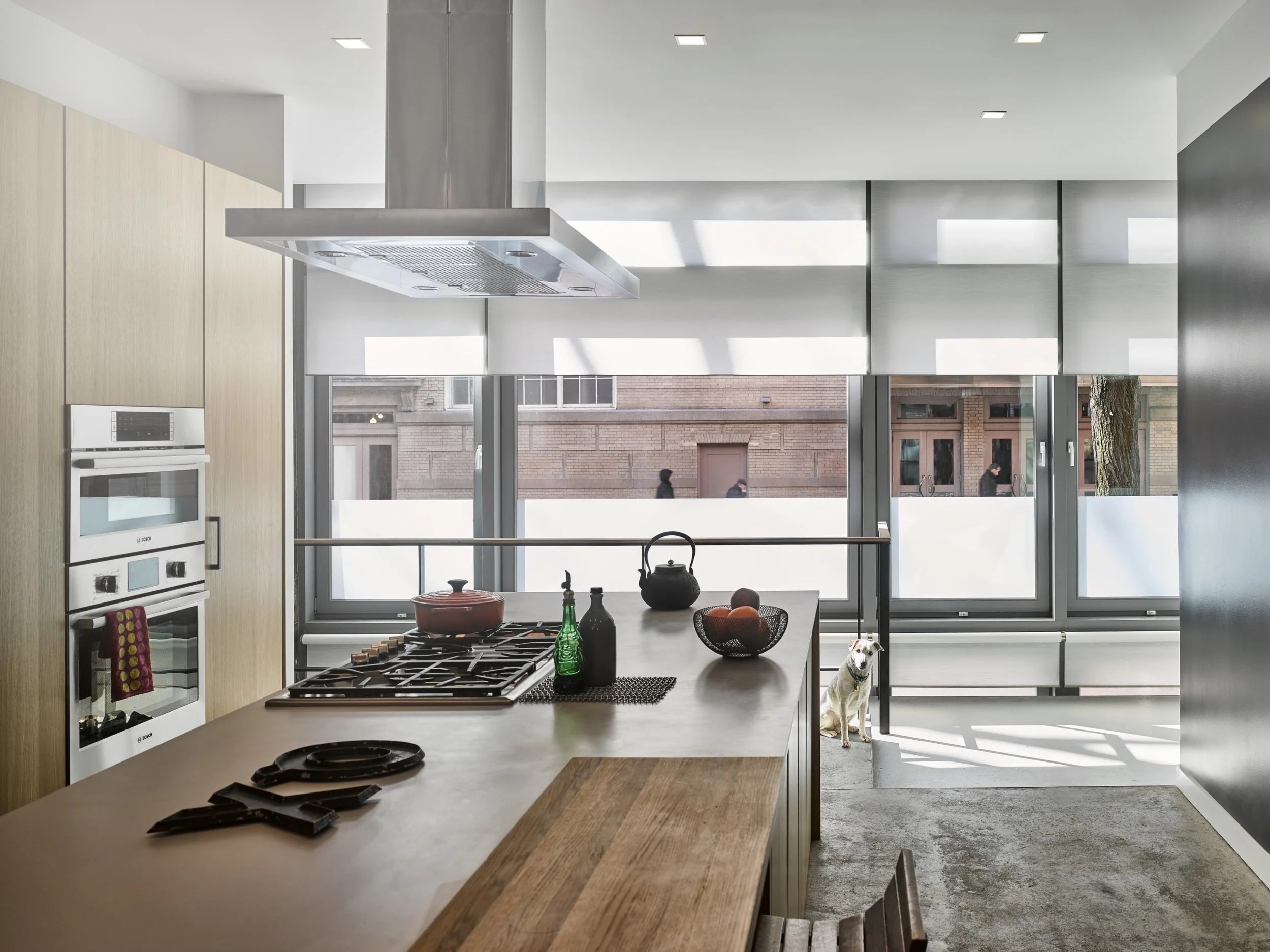 A modern city-view kitchen featuring Lutron motorized roller shades that filter daylight while maintaining privacy across expansive windows. The streamlined island cooktop with range hood sits at the center, paired with integrated built-in ovens and