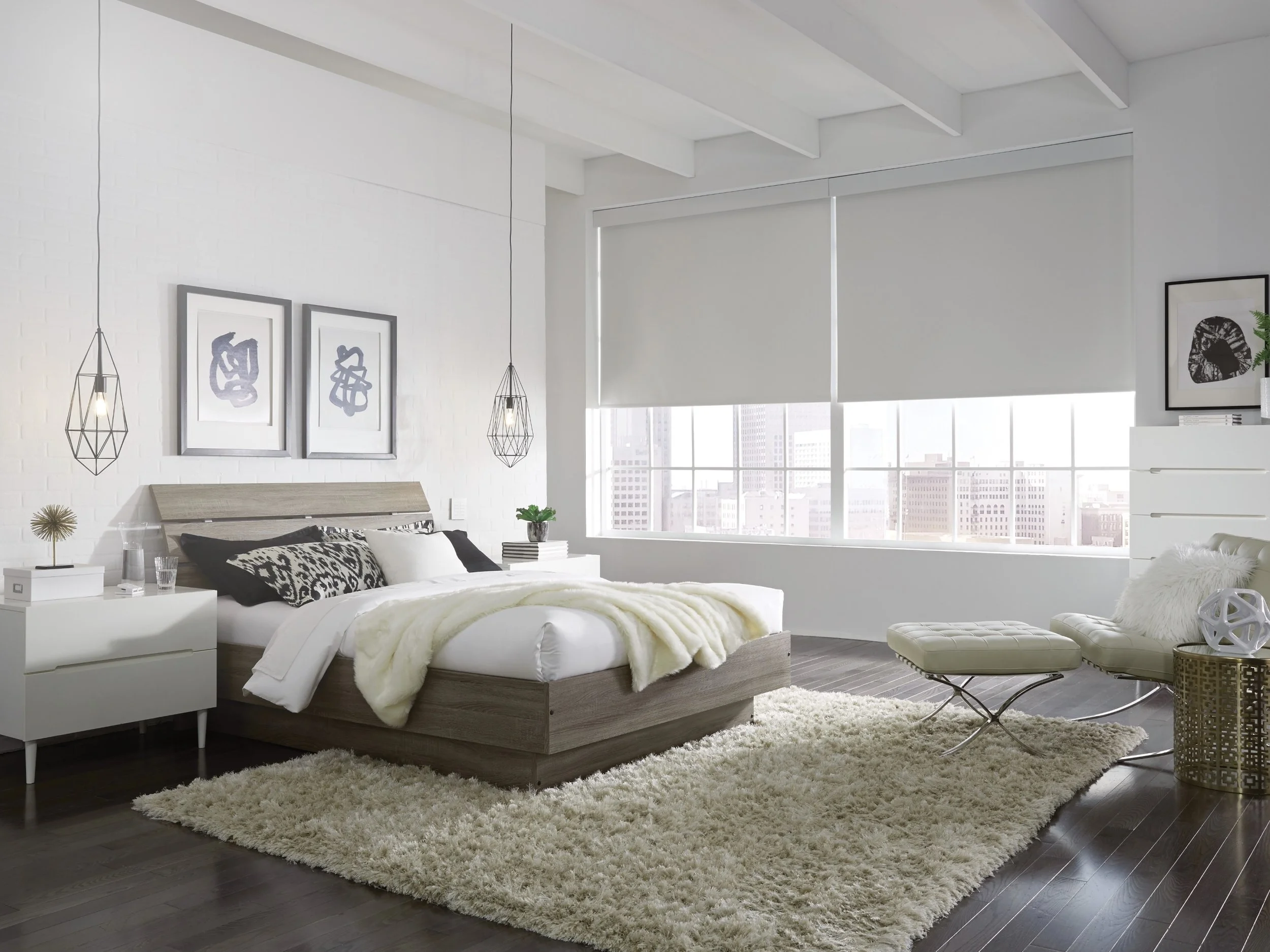 A modern bedroom featuring Lutron QS roller shades integrated with HomeWorks QSX lighting and shading control. The room showcases clean architectural lines with white walls, dark wood flooring, and exposed ceiling beams, softened by a plush shag rug