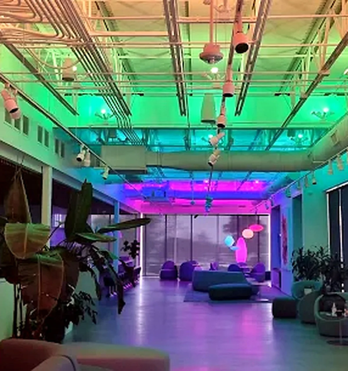 An indoor lounge area with colorful LED lighting on the ceiling, featuring green, pink, and purple hues. The space includes lounge chairs, potted plants, and large windows, illuminated by decorative hanging lamps.